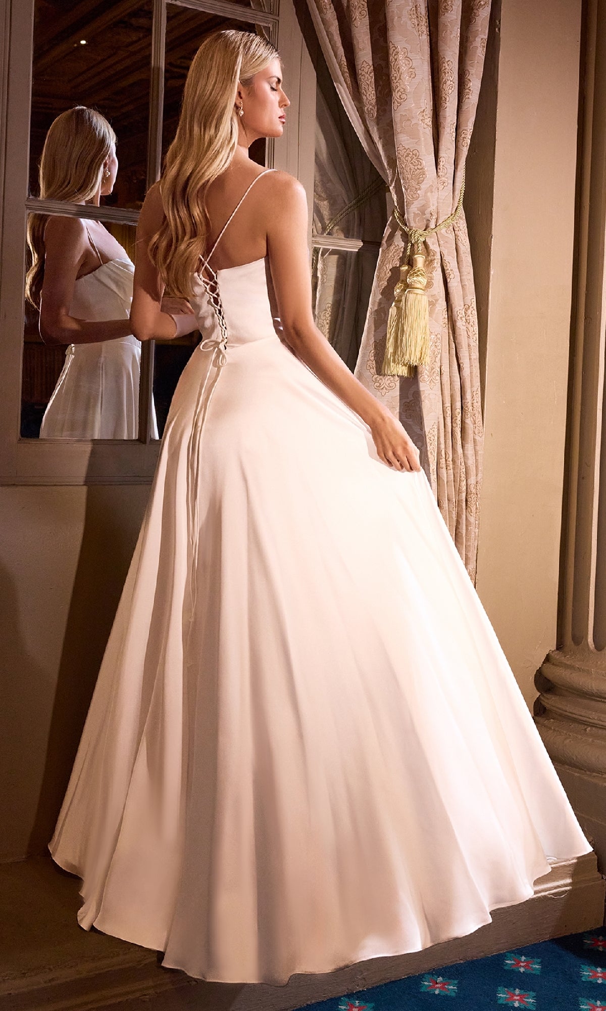 Wearing the Ladivine 7481W Lace-Up Long White Wedding Dress, a woman stands near a window, her reflection softly visible in the glass.