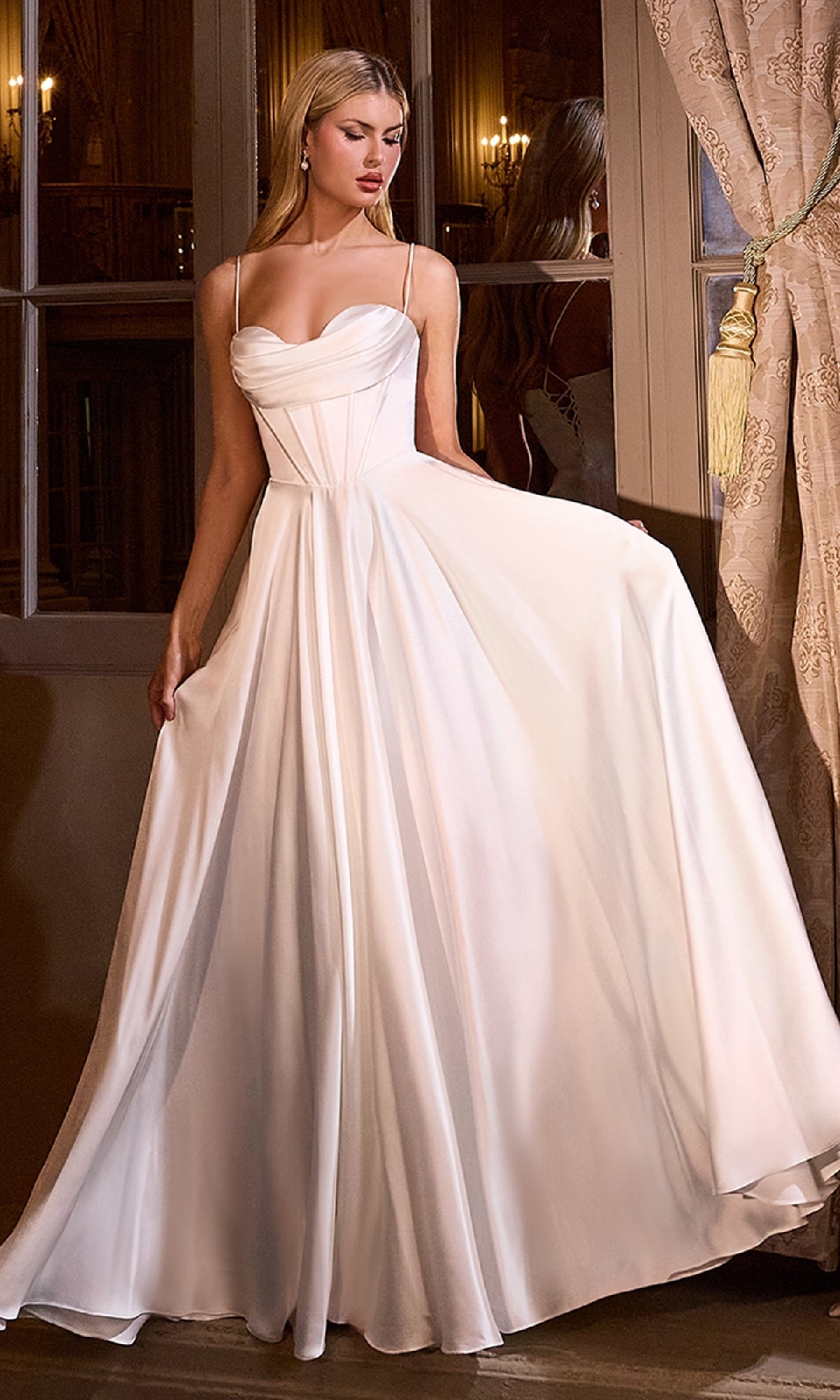 A woman in the Lace-Up Long White Wedding Dress: Ladivine 7481W stands indoors by a window, holding out part of her elegant, floor-length off-white gown with thin straps.