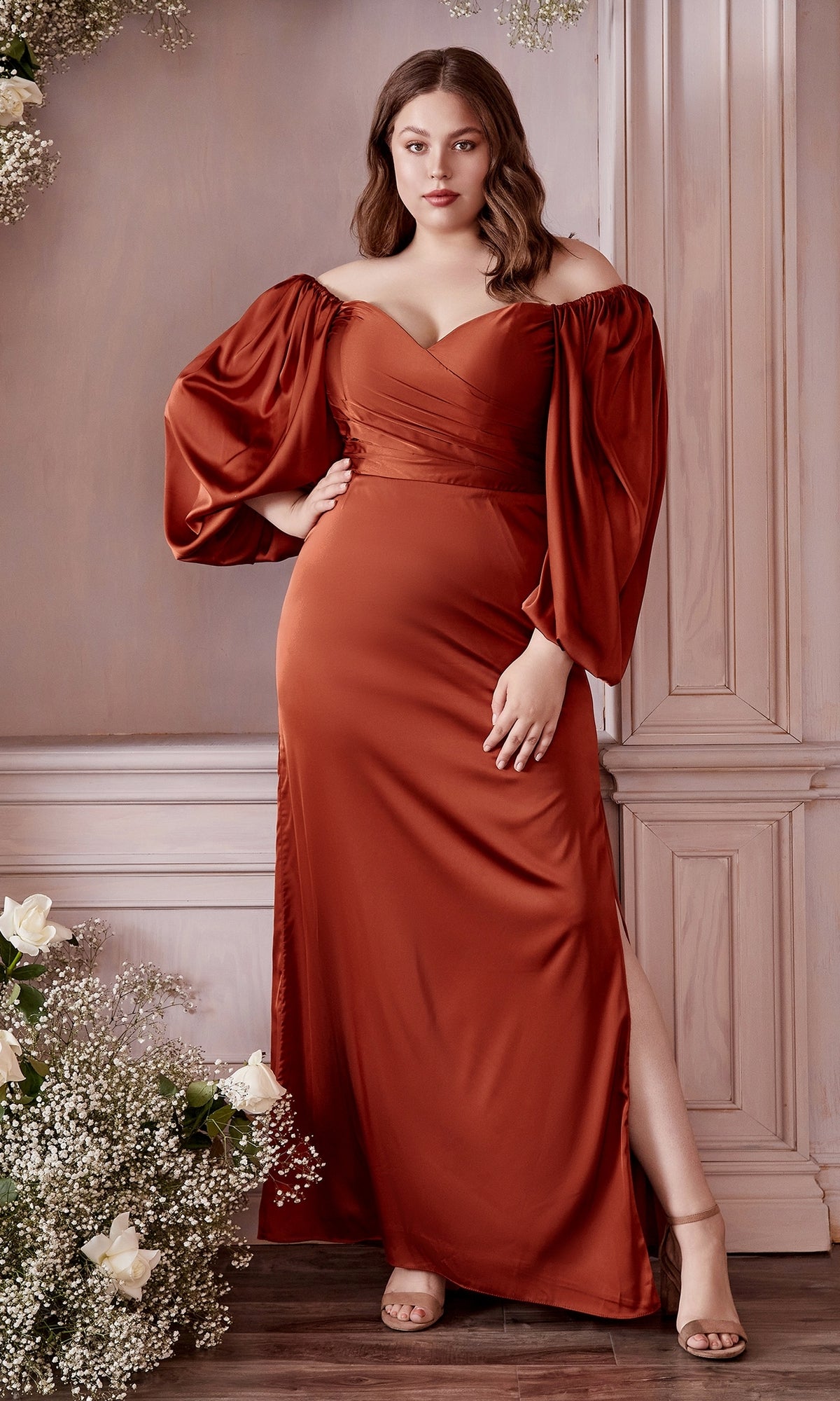 A woman in the Ladivine 7482C Plus Long Sleeve Long Prom Dress, featuring rust color and off-the-shoulder design, stands indoors by wooden paneling and white flowers.