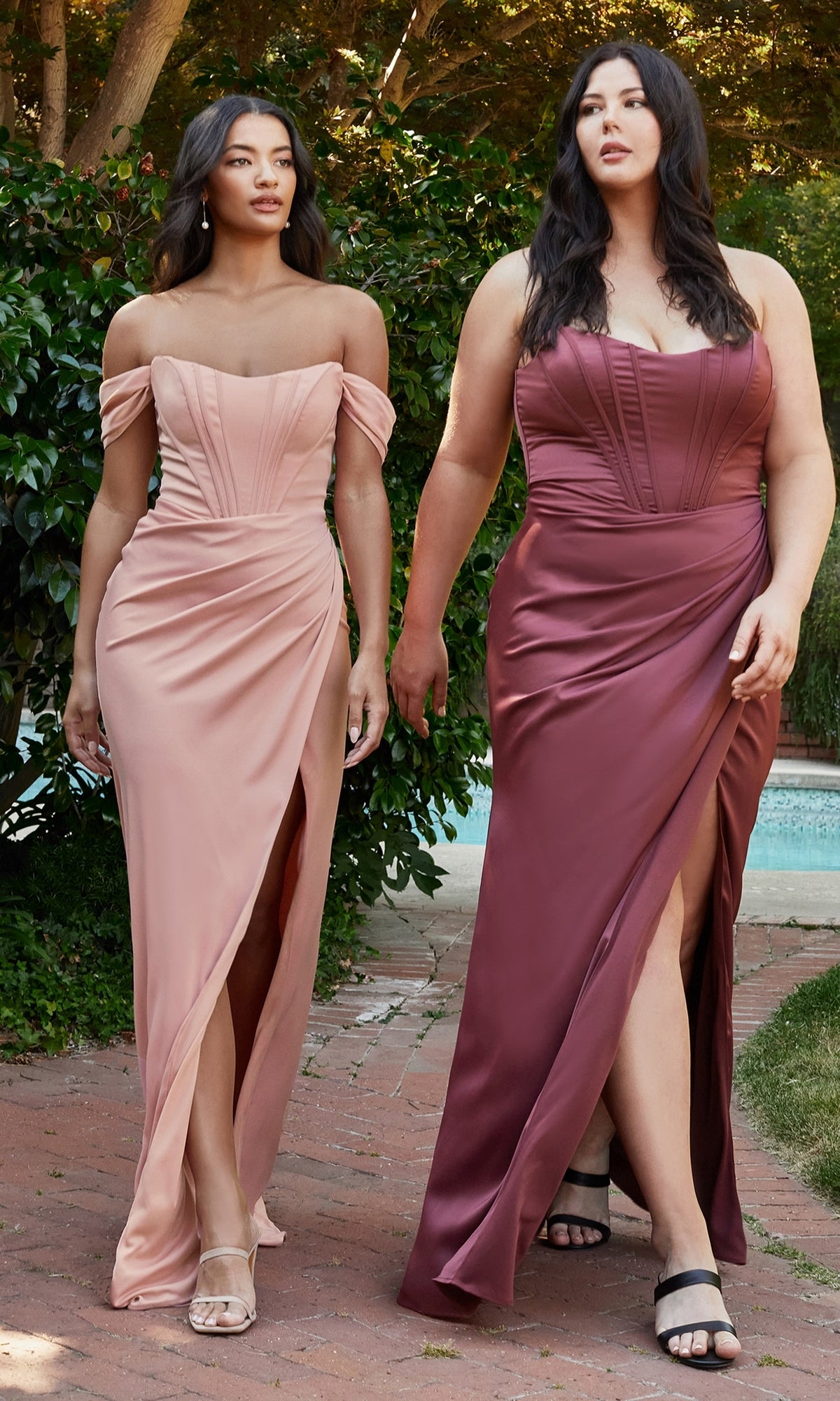 Two women in Corset Off-the-Shoulder Long Prom Dress 7484, featuring thigh-high slits, walk outdoors on a brick path bordered by lush greenery and a shimmering pool.