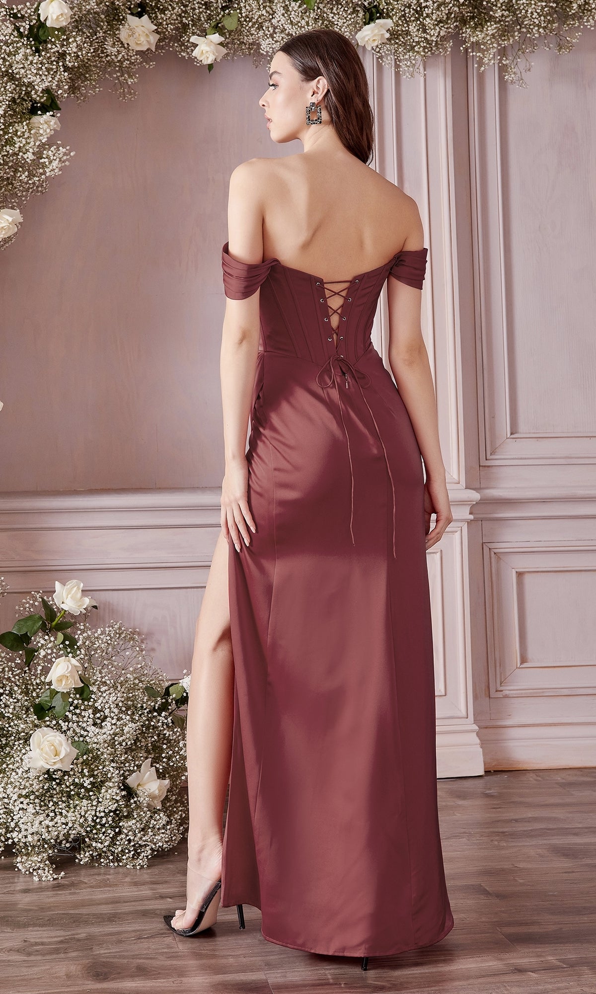 A woman stands on a wooden floor in the Off-the-Shoulder Long Plus-Size Prom Dress 7484C—a mauve gown with lace-up back and thigh-high slit—surrounded by white floral arrangements against a paneled wall.