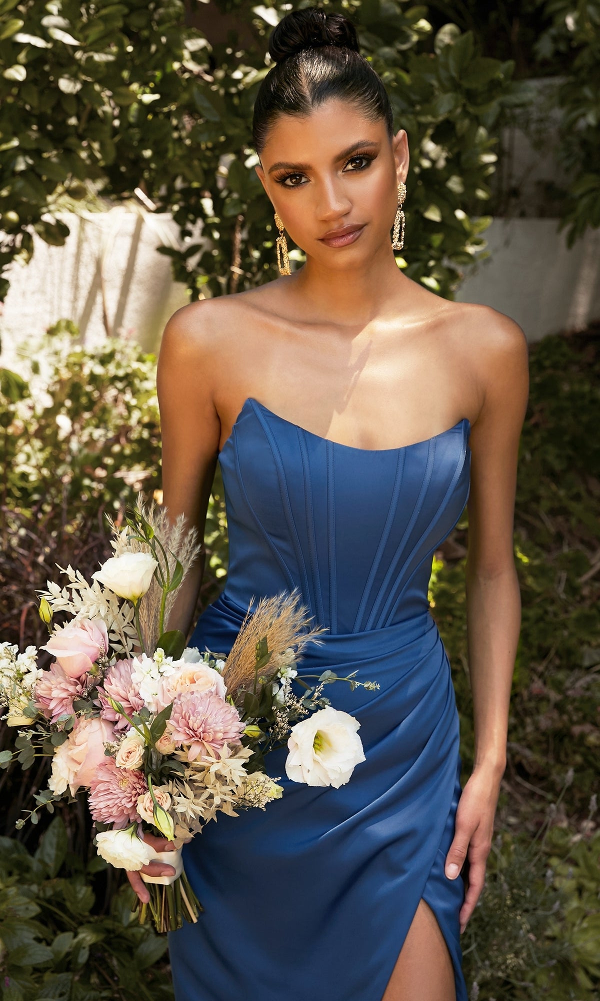Woman in a Corset Off-the-Shoulder Long Prom Dress 7484 holds a bouquet of flowers, standing outdoors against a lush green backdrop.