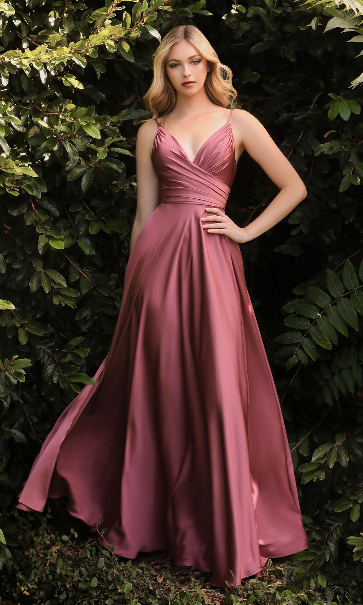 A woman stands outdoors before green foliage, wearing the Classic Long Satin Formal Prom Dress: Ladivine 7485—a floor-length, sleeveless rose satin gown with a V-neckline and pleated bodice.