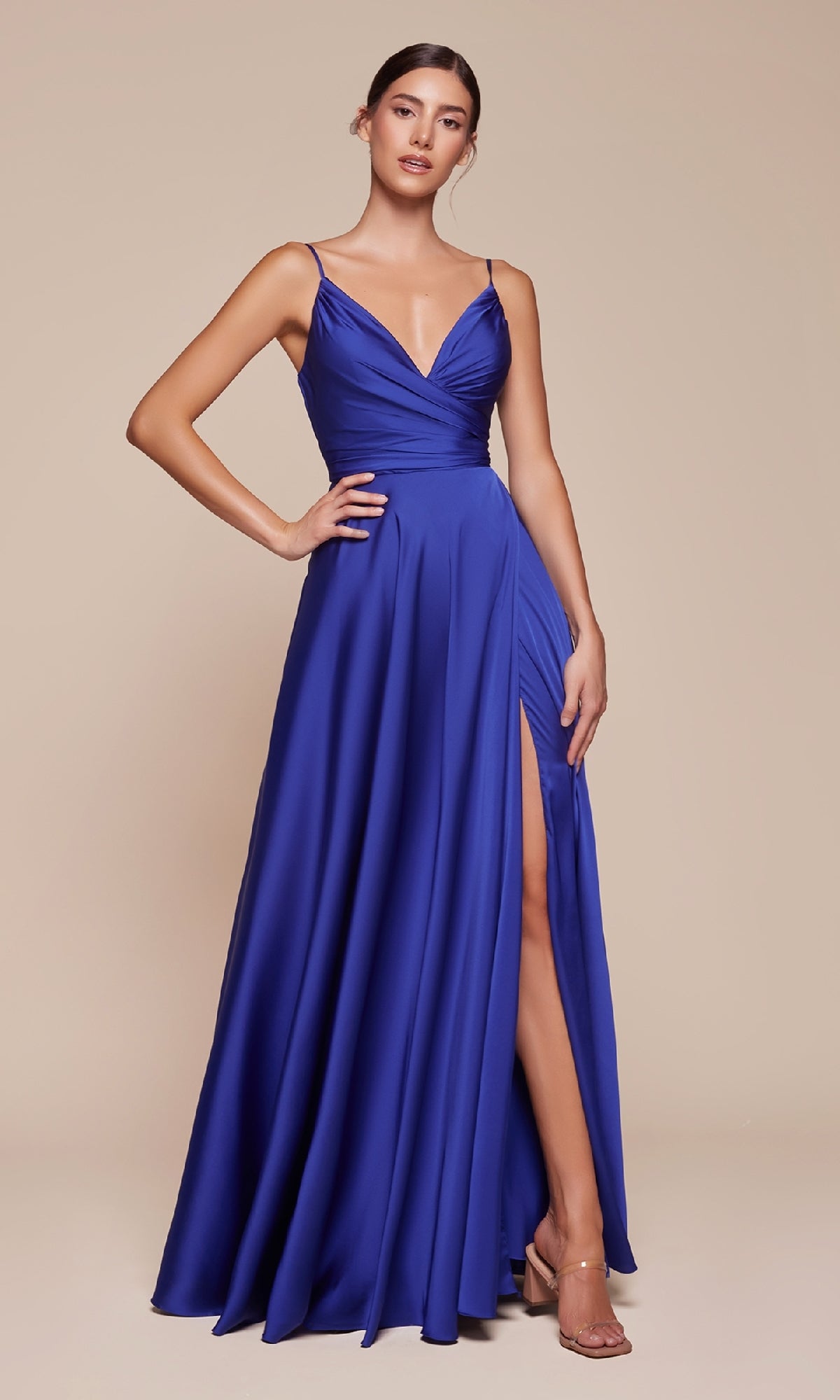 A woman in the Classic A-Line Satin Prom Dress: Ladivine 7485-1, a sleeveless royal blue gown with a deep V-neck, gathered bodice, high slit, and spaghetti straps, stands against a beige background.