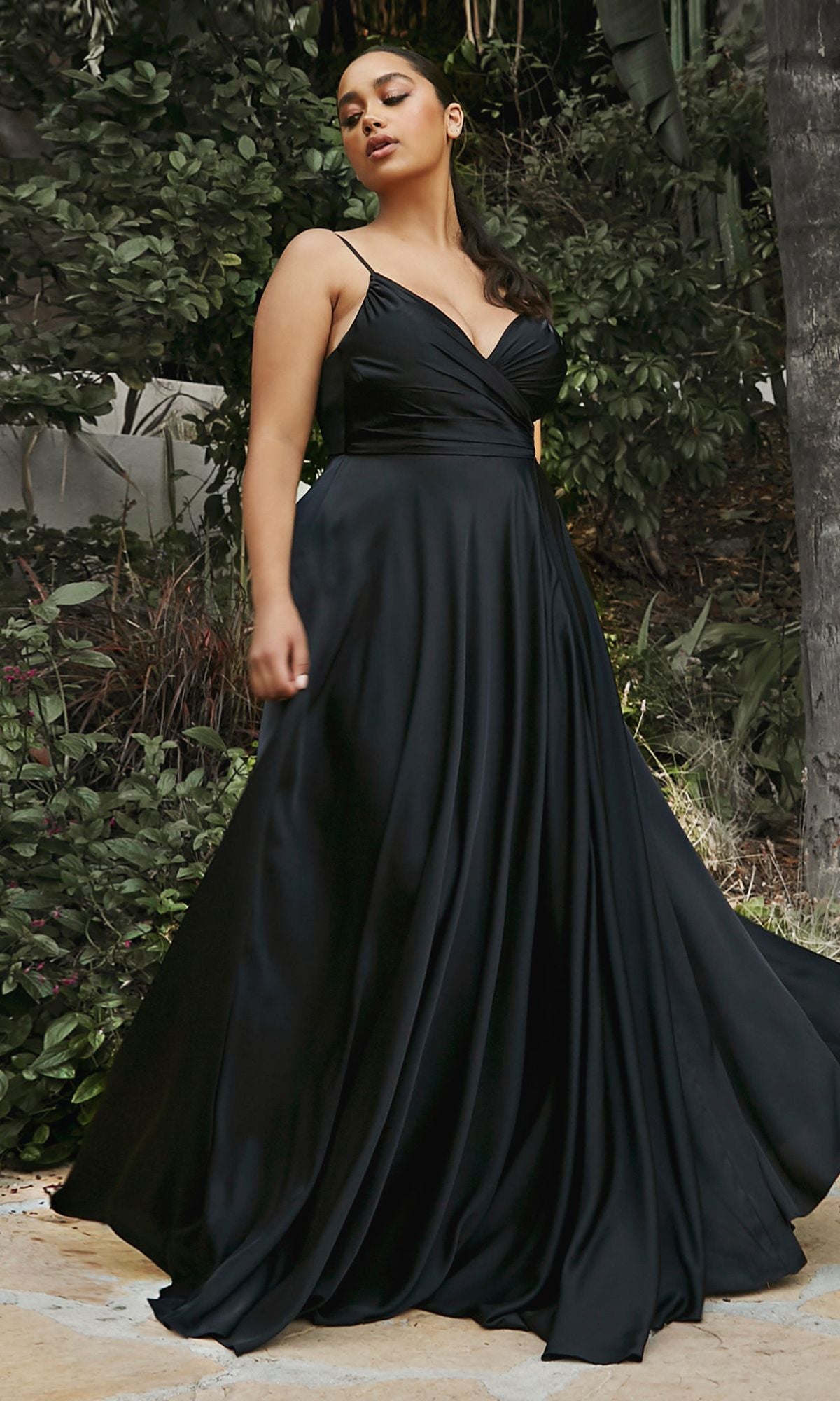 A woman stands outdoors in a Classic Long Satin Formal Prom Dress: Ladivine 7485, a floor-length black gown with a deep V-neckline and A-line skirt, surrounded by greenery.