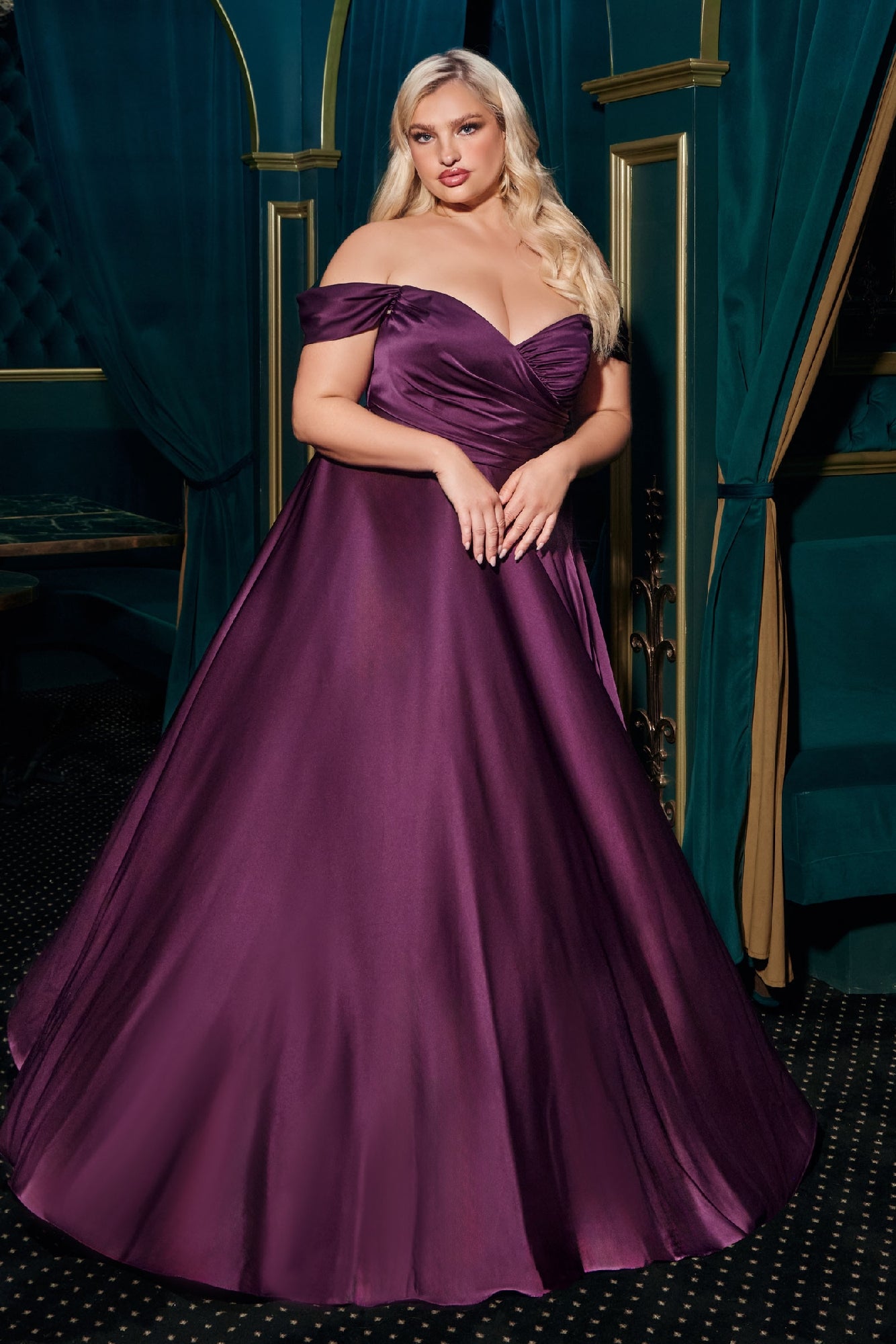 A woman showcases the Empire-Waist Plus-Size Long Prom Dress 7493C in an elegant room with teal drapes and gold accents, wearing a floor-length, off-the-shoulder purple gown.