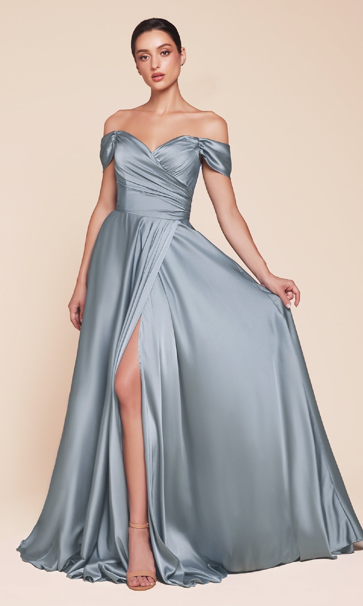A woman models the Ladivine 7493-1, an off-the-shoulder long prom dress in silver satin with a thigh-high slit, standing against a beige background.