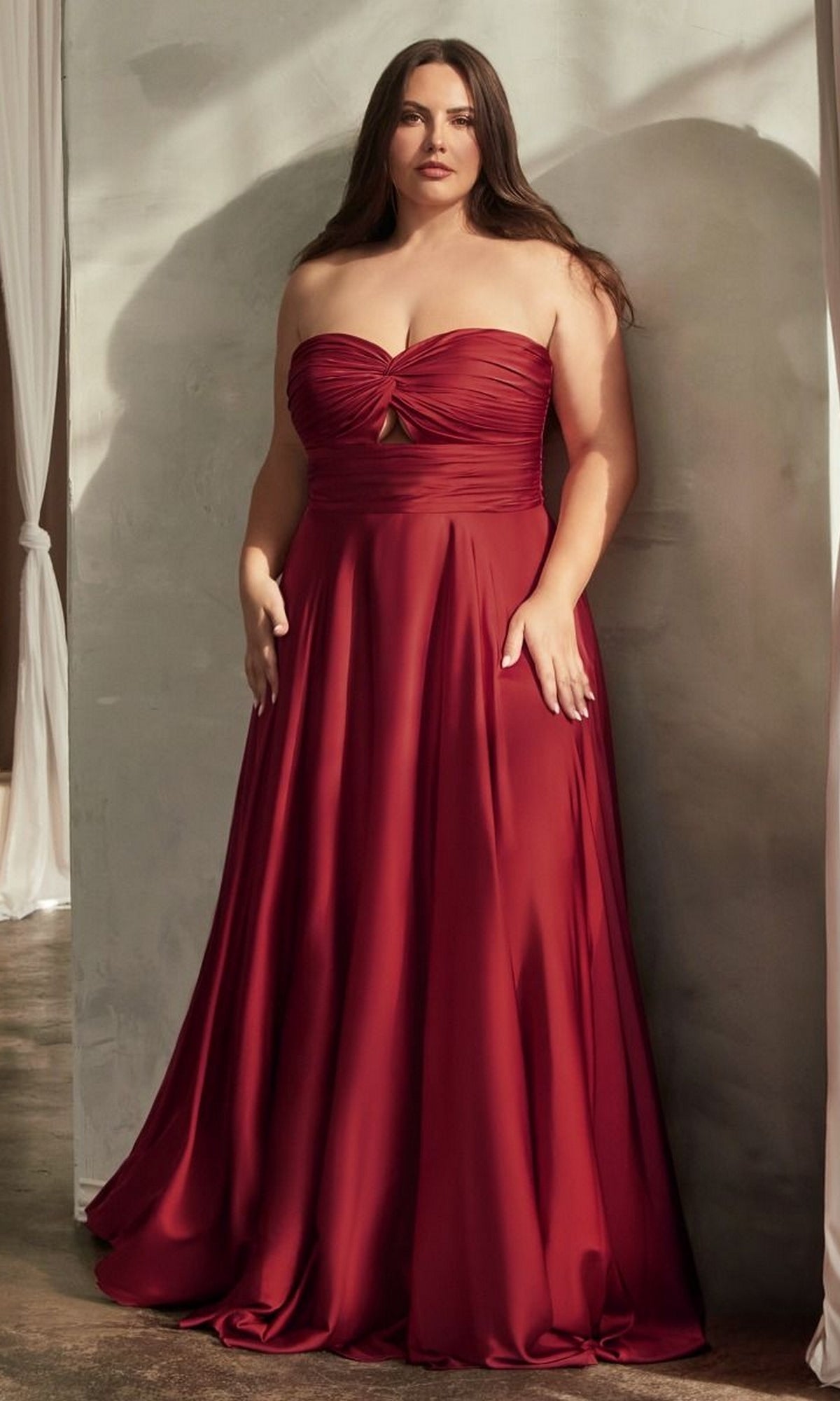 A woman stands against a light wall in the Plus-Size Strapless Sweetheart Prom Dress 7496C—a floor-length red gown with a ruched bodice and small keyhole cutout.