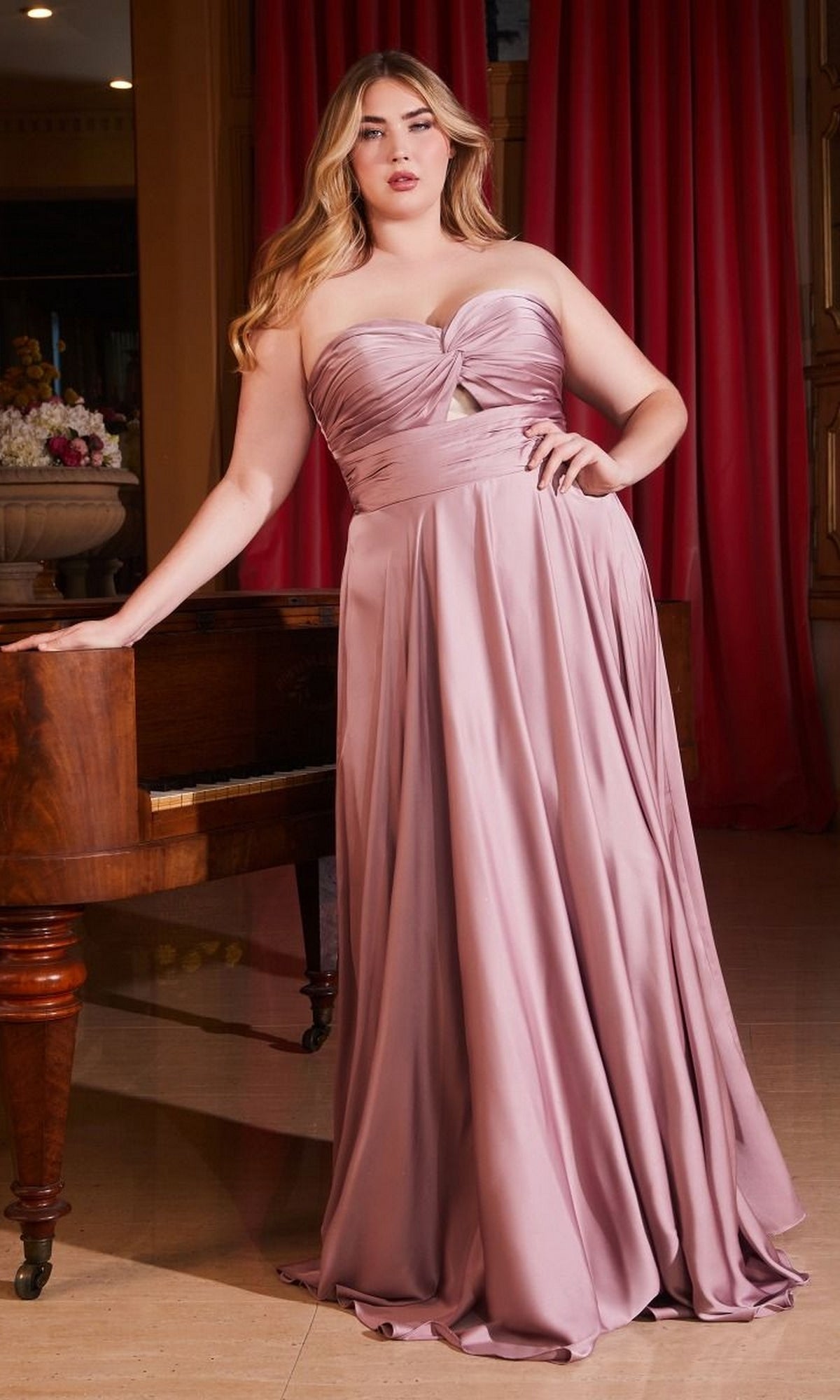 A woman in a Plus-Size Strapless Sweetheart Prom Dress 7496C in dark mauve stands by a wooden piano framed by elegant red curtains, her floor-length gown making an affordable and stylish evening dress option.