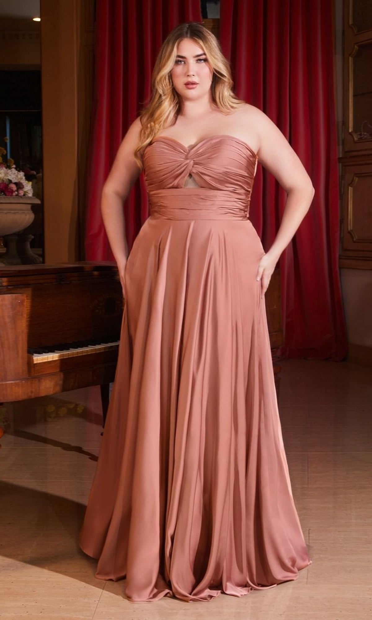 A woman stands indoors in the Plus-Size Strapless Sweetheart Prom Dress 7496C, a rose-colored, floor-length gown with a gathered bodice and side pockets. A piano and red curtains are in the background. This elegant gown is an affordable evening dress choice.
