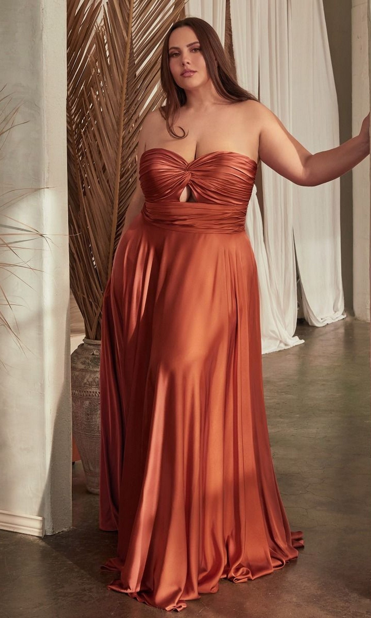 A woman in the Plus-Size Strapless Sweetheart Prom Dress 7496C—a burnt orange satin gown—stands indoors by neutral-toned decor and draped white curtains.