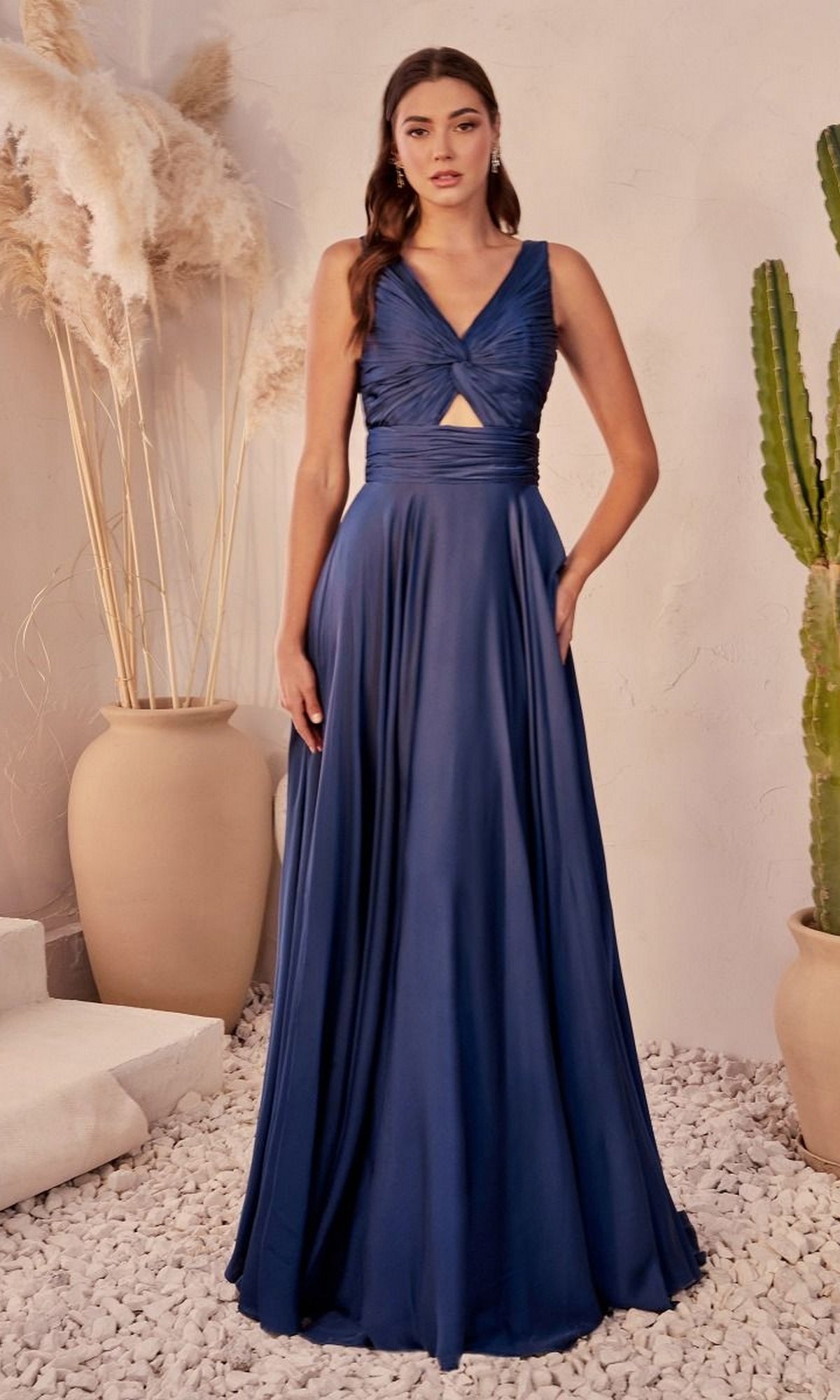 Indoors, a woman models the Twist-Front Long A-Line Cut-Out Prom Dress 7497, featuring a sleeveless design, floor-length navy blue skirt, and front keyhole detail. She stands among large vases and decorative plants.