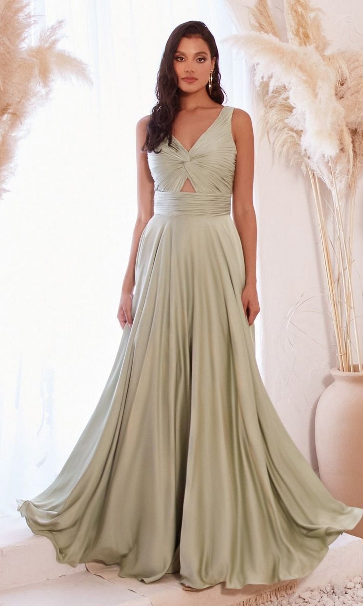 A woman stands indoors among tall pampas grass, wearing the Twist-Front Long A-Line Cut-Out Prom Dress 7497 in sage green, featuring a stylish twist-front cutout and a flowing skirt.