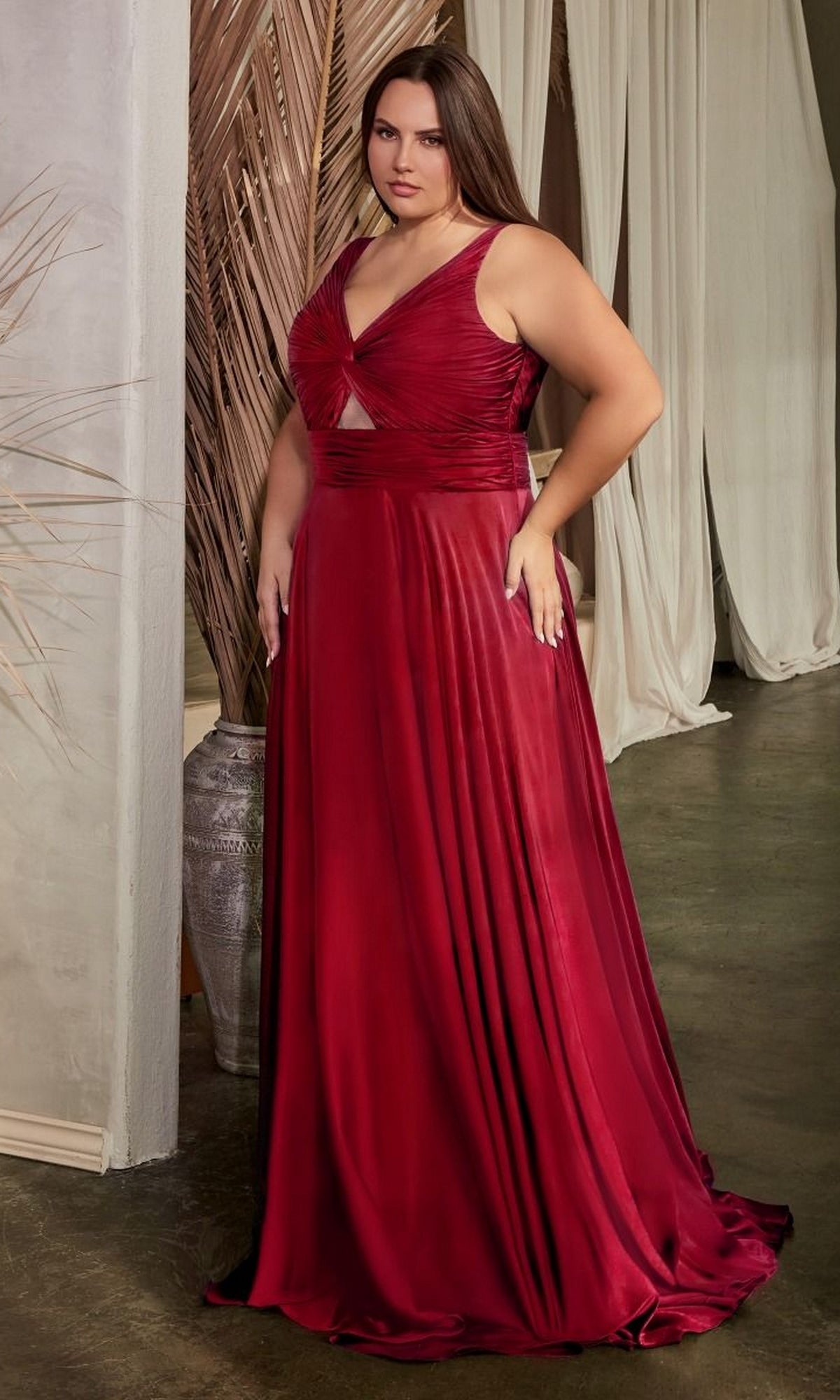 A woman stands indoors near draped curtains and decorative dried plants, wearing the Twist-Front Long Plus-Size Prom Dress 7497C—a sleeveless, floor-length red formal gown.