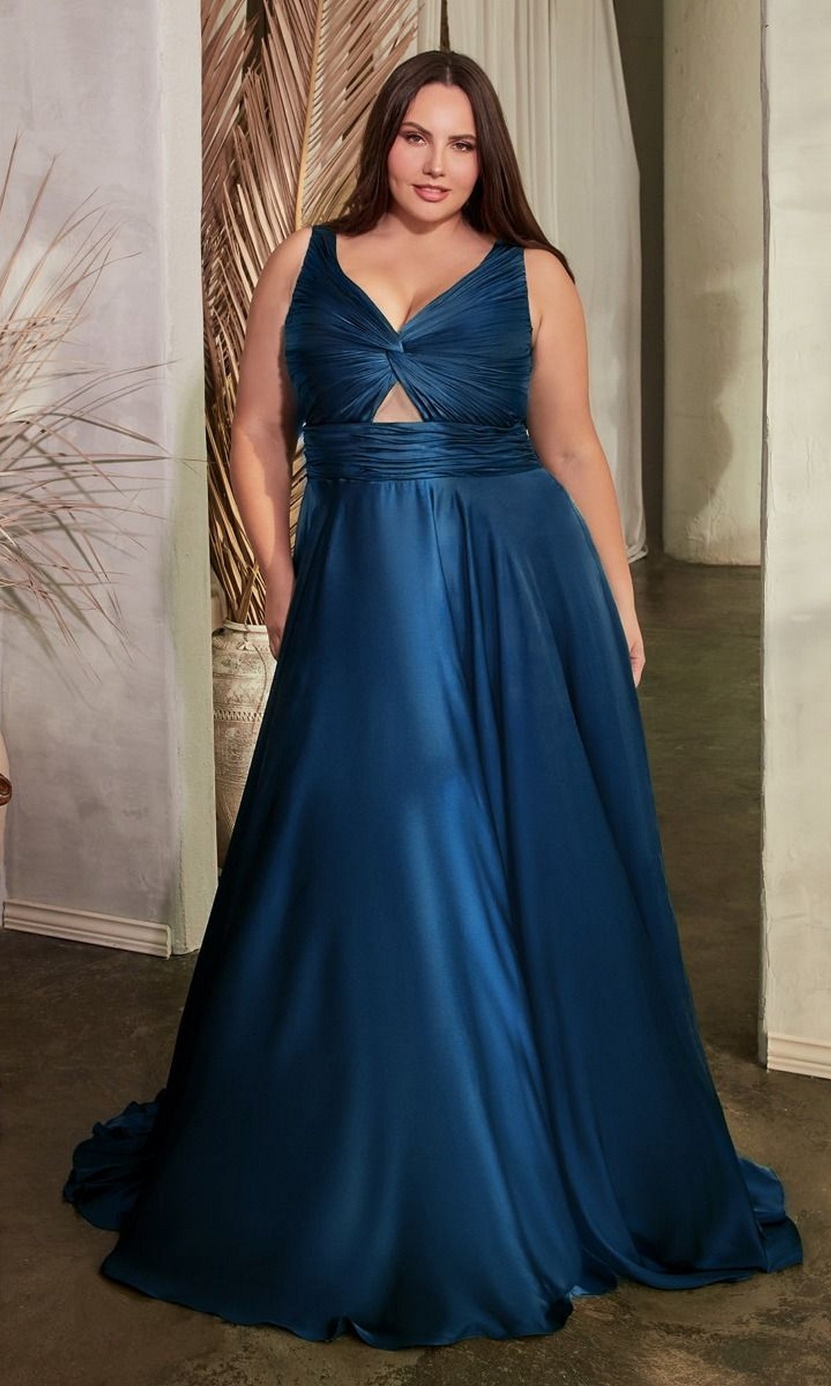 A woman wearing the Twist-Front Long Plus-Size Prom Dress 7497C—a sleeveless, deep blue formal gown with a v-neckline and front cutout—stands indoors on a concrete floor.