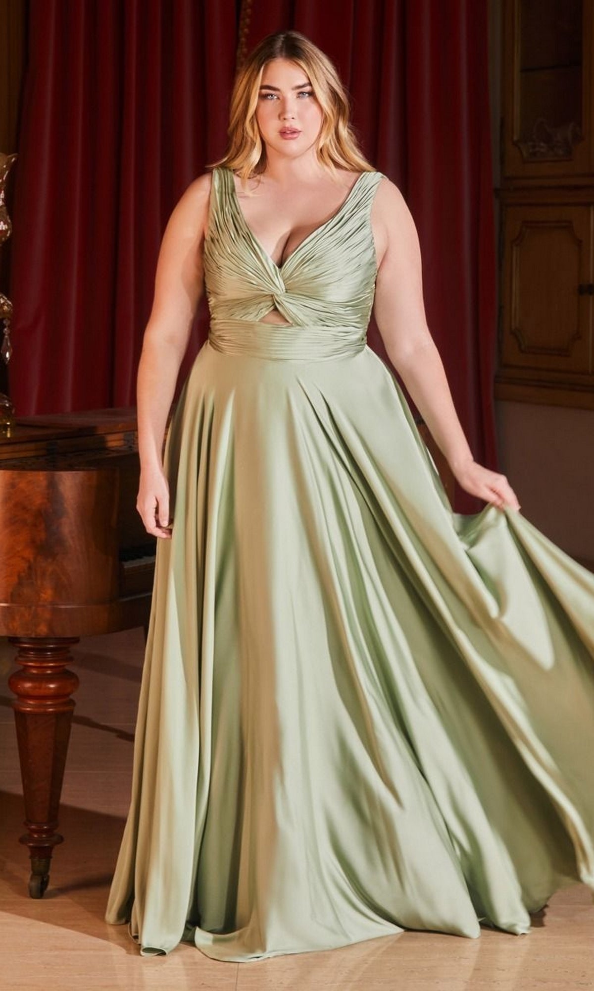 A woman in the Twist-Front Long Plus-Size Prom Dress 7497C, light green with a twisted bodice, stands indoors holding her gown. Red curtains and a wooden piano create an elegant backdrop.