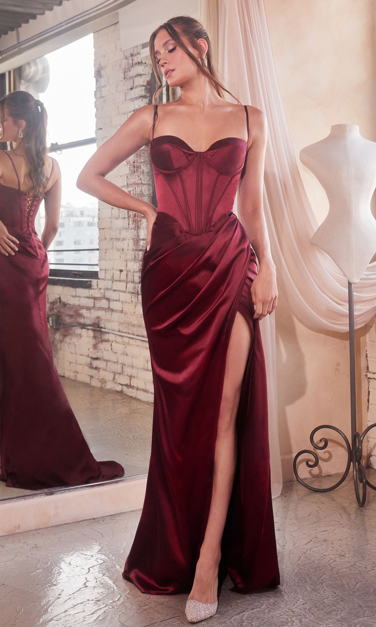 A woman wearing the Bustier-Bodice Draped Long Prom Dress 7498 in burgundy satin, featuring a bustier bodice and thigh-high slit, stands beside a mirror and dress form in a softly lit room.