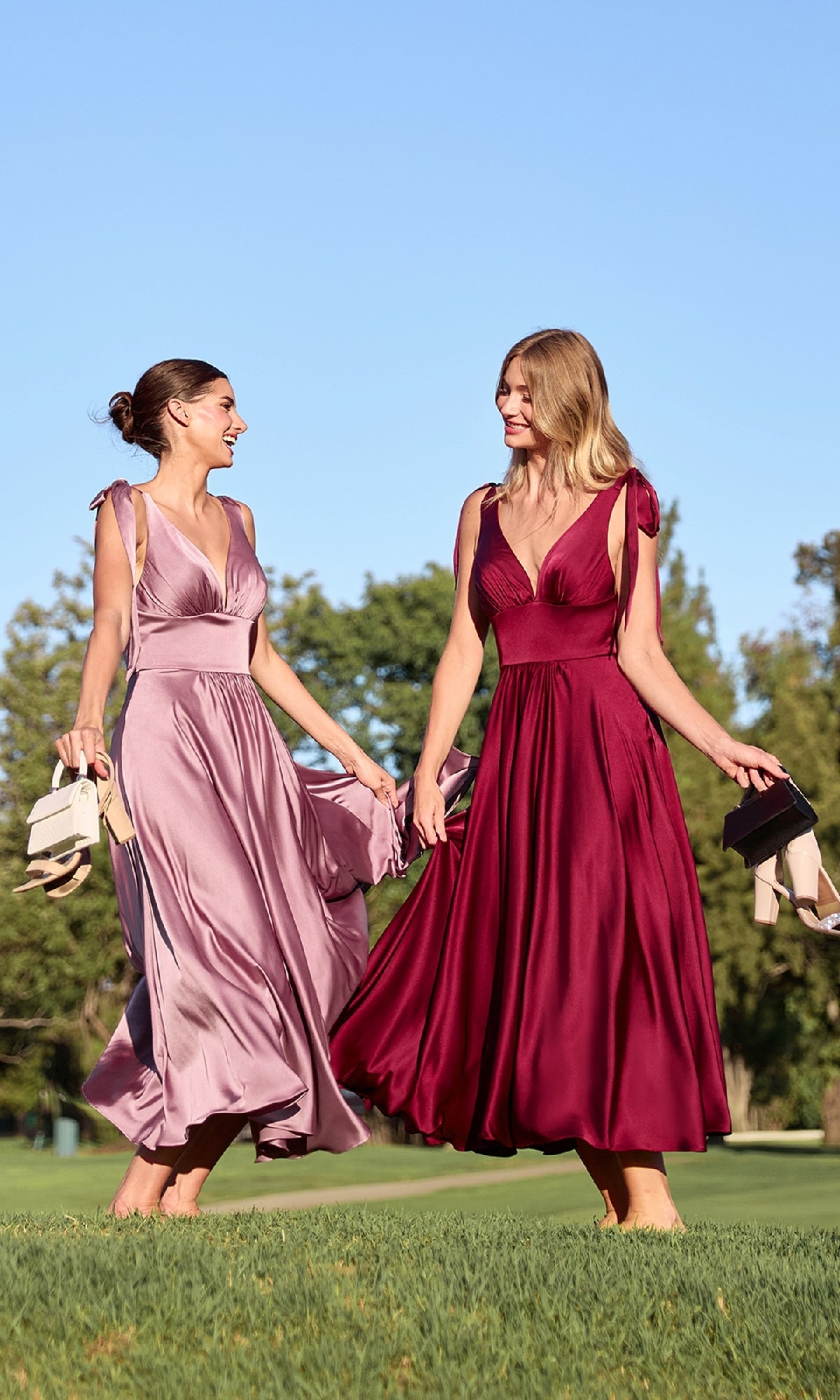 Two women in Ladivine 7499 tea-length wedding-guest dresses—one lavender, one burgundy—walk barefoot on grass, holding their shoes, with trees and a clear blue sky in the background.