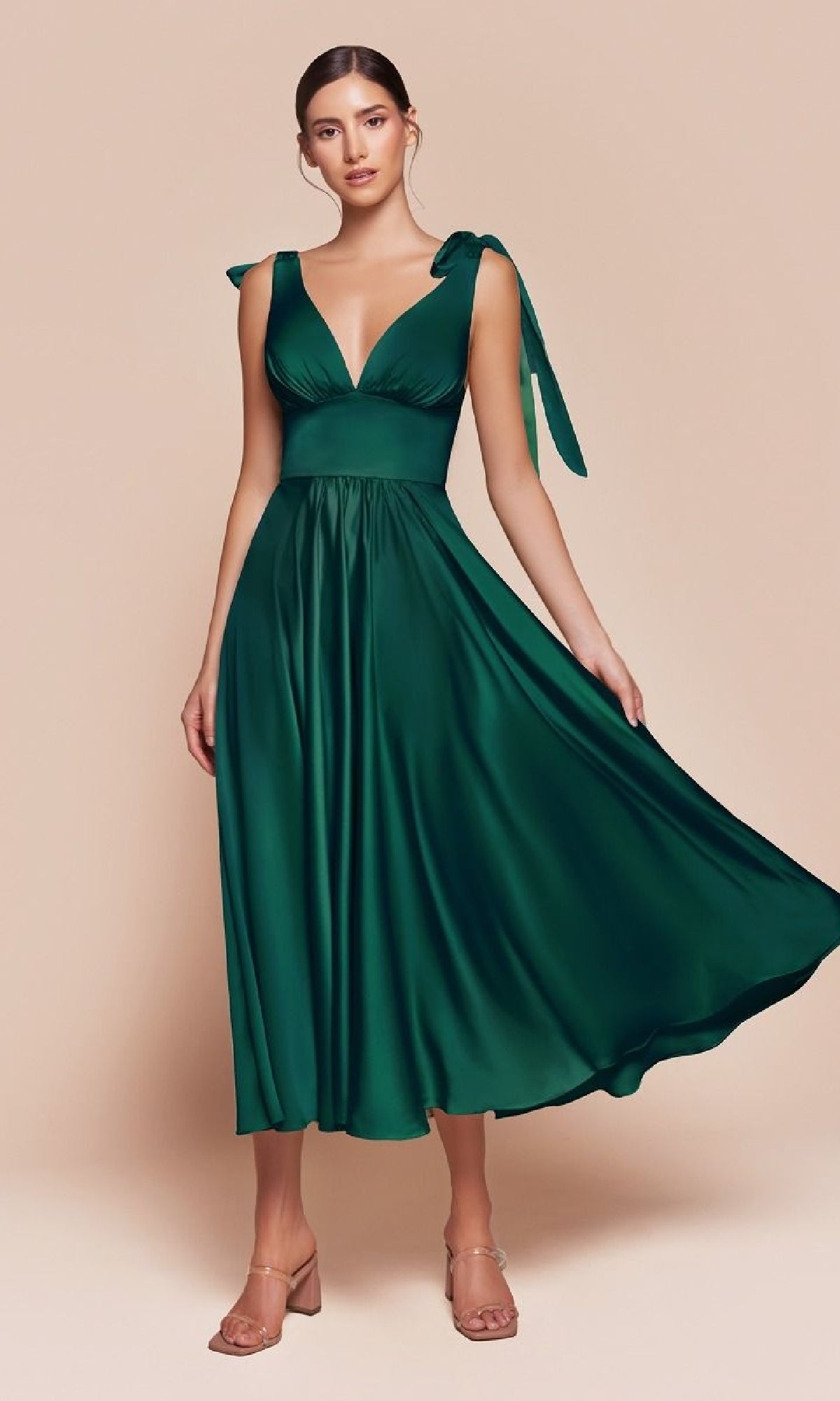 A woman in the Ladivine 7499 tea-length wedding-guest dress, featuring a sleeveless deep green satin design with a fitted waist and flowing skirt, stands against a plain beige background.