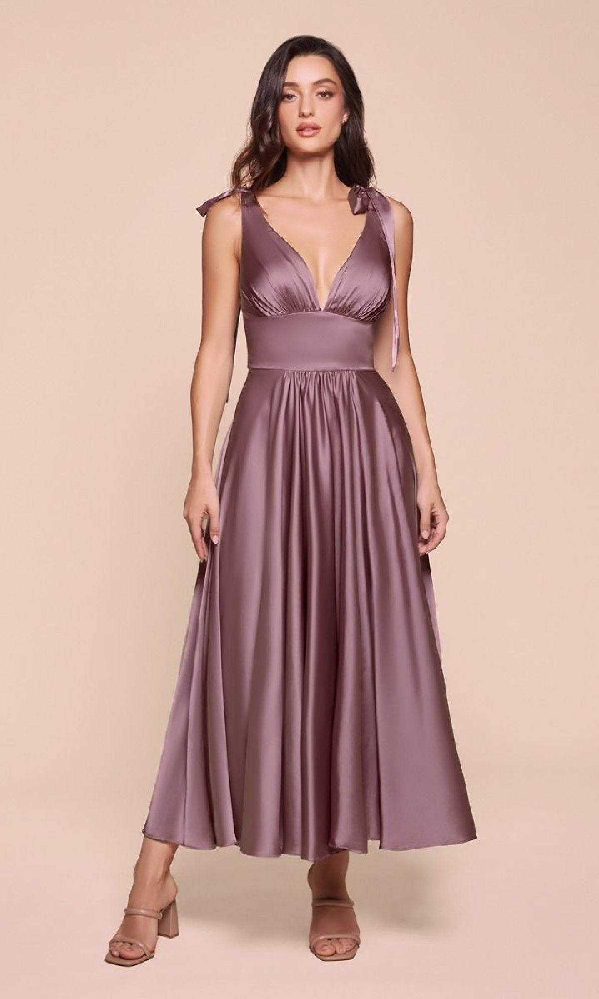 A woman wears the Ladivine 7499, a sleeveless mauve satin tea-length wedding-guest dress with V-neckline, tied shoulder straps, fitted waist, and flowing ankle-length skirt, paired with open-toed heels.