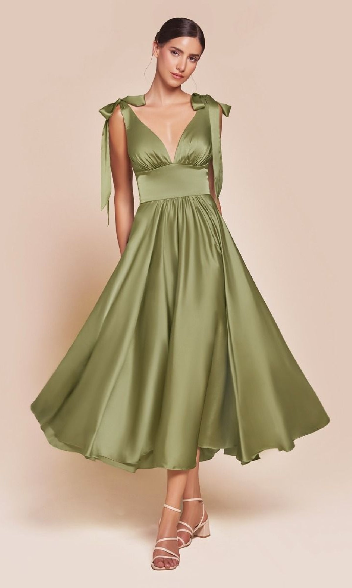 A woman wears the Ladivine 7499 tea-length wedding-guest dress, an olive green, sleeveless a-line midi with a deep V-neck and bow-tied shoulder straps, paired with strappy beige sandals.
