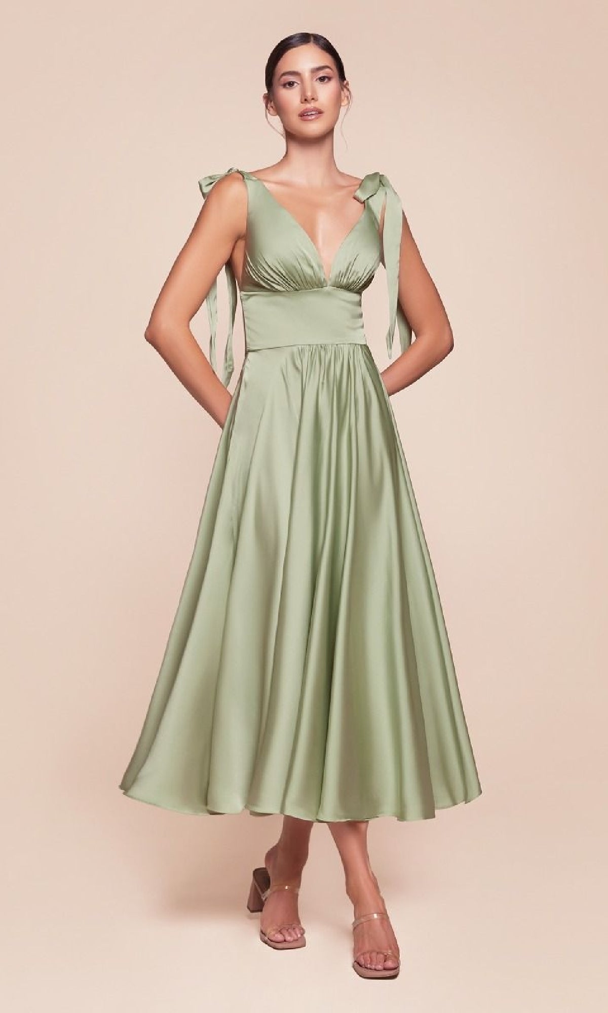 Woman wearing the Ladivine 7499, a light green, tea-length wedding-guest dress featuring bow shoulder straps and a flared skirt, stands against a plain beige background.