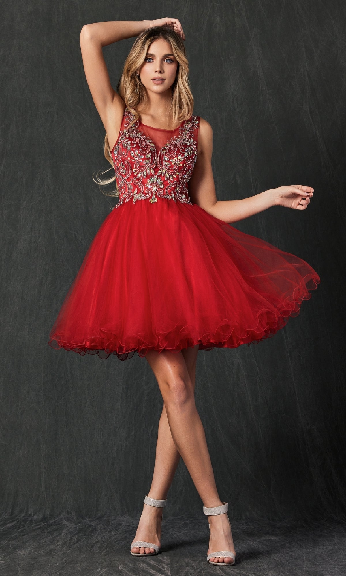 A woman wears the Short Hoco Dress: Juliet 749—a sleeveless red homecoming dress with an embellished bodice, intricate silver embroidery, and an elegant A-line tulle skirt—standing against a dark background.