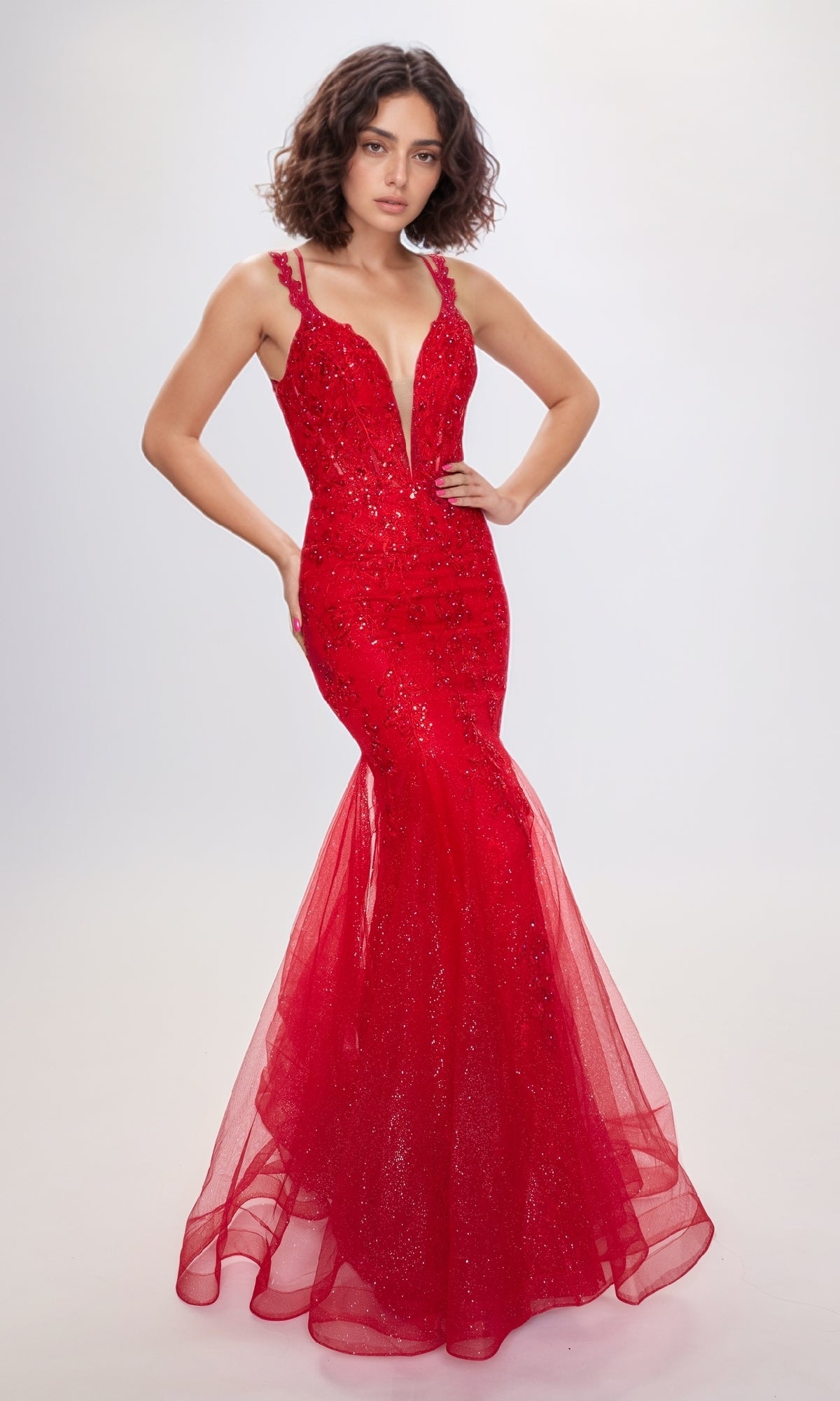 A woman wears the Nina Canacci 7600 Lace Mermaid Prom Dress, a sleeveless, form-fitting red gown with a deep V neckline and sparkling embellishments, standing against a plain light background.