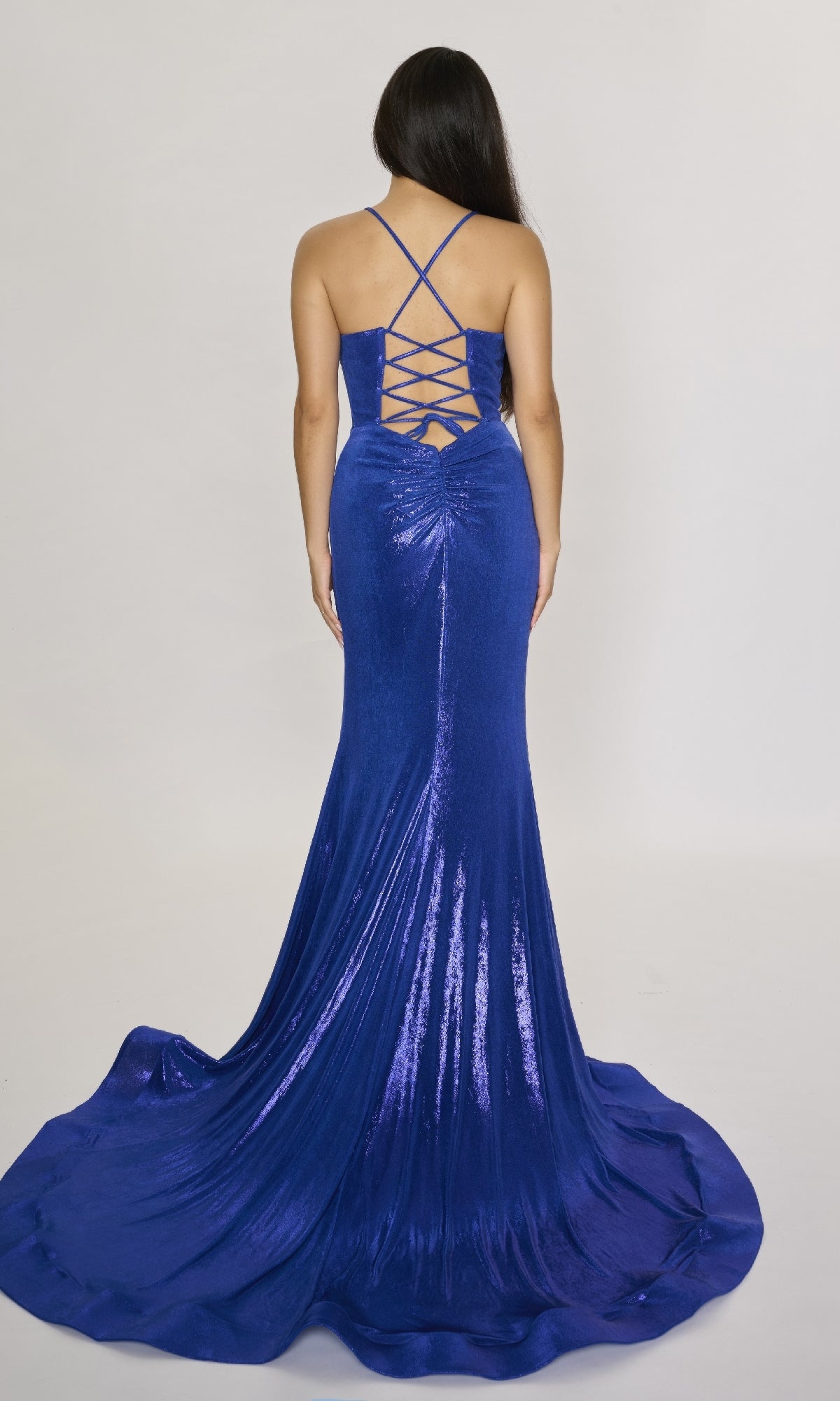A woman in the Nina Canacci 7604 stands with her back to the camera, showcasing its metallic cowl bodice, lace-up open back, and shimmering long blue gown against a plain background.
