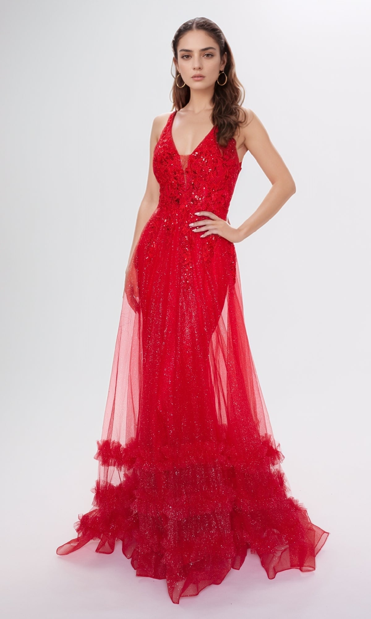 A woman wears the Nina Canacci 7607, a sleeveless floor-length red sequin formal dress with a deep V-neckline, fitted silhouette, and tulle ruffles at the hem, standing against a plain background.