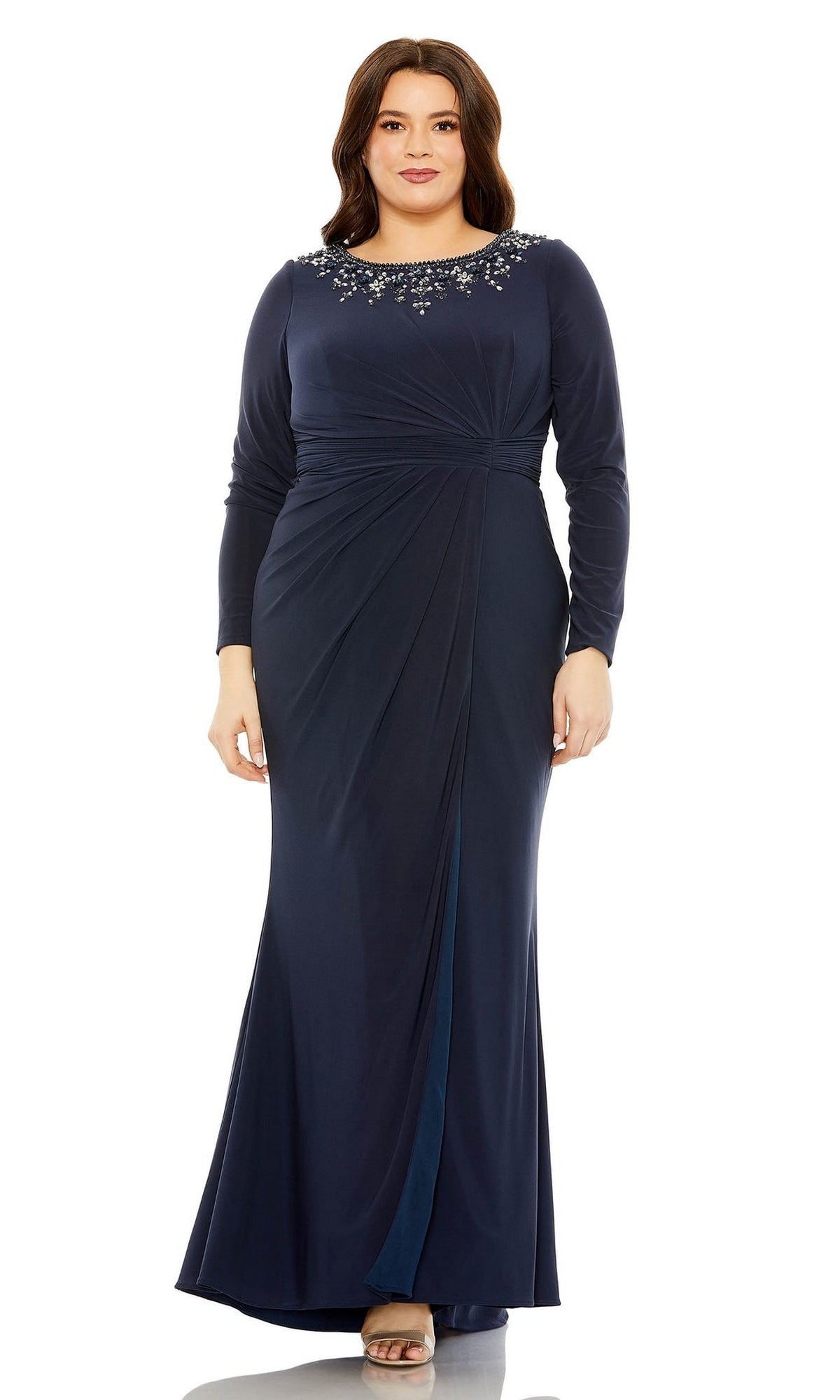 A woman stands facing forward in the Mac Duggal Long Plus-Size Prom Dress 77023—a navy blue, floor-length gown featuring long sleeves, a gathered waist, and an embellished neckline.