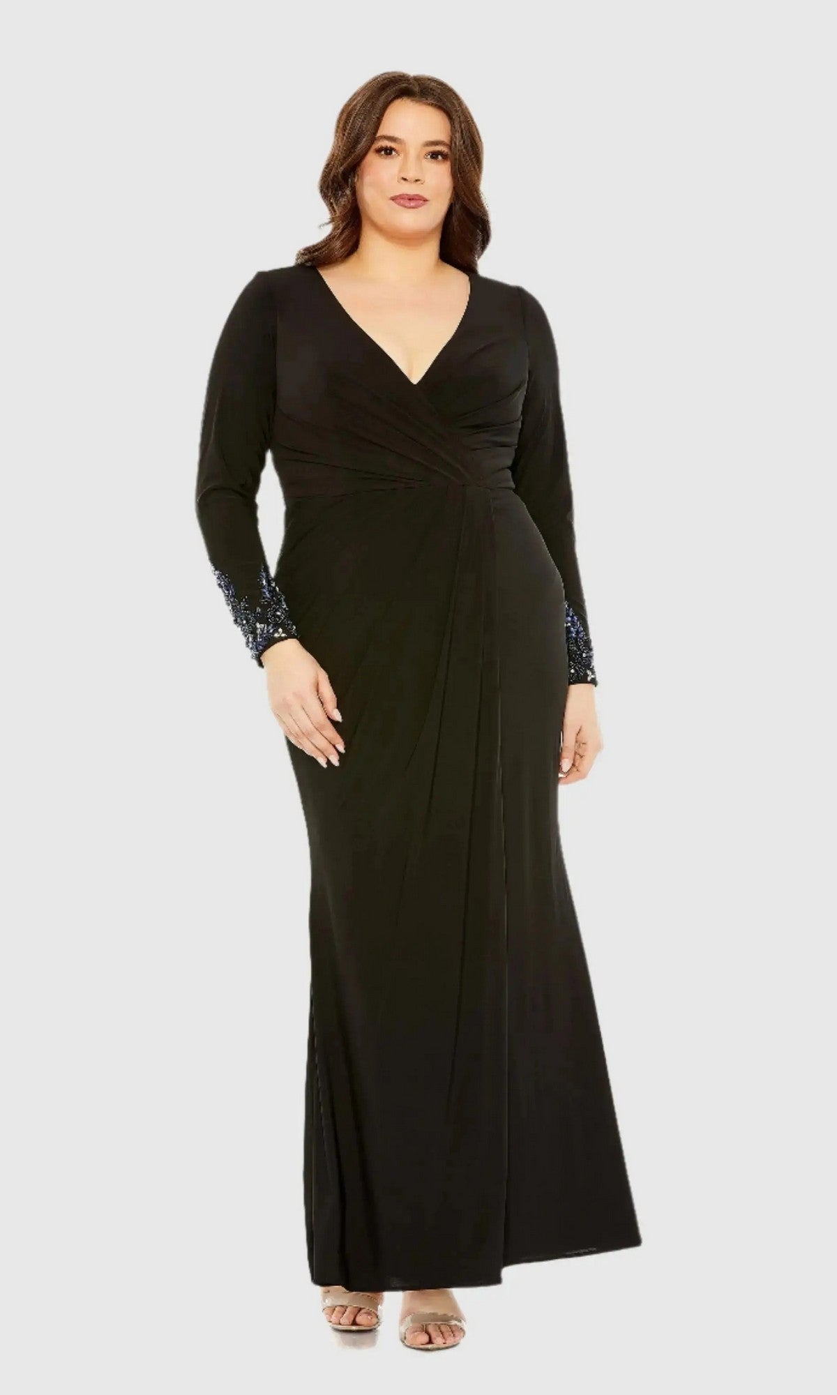 A woman models the Long Plus-Size Formal Dress: Mac Duggal 77024 in black, featuring a long wrap silhouette with a V-neckline, long sleeves, and embellished cuffs. She stands facing forward against a plain light gray background, showcasing this elegant plus-size formal gown.