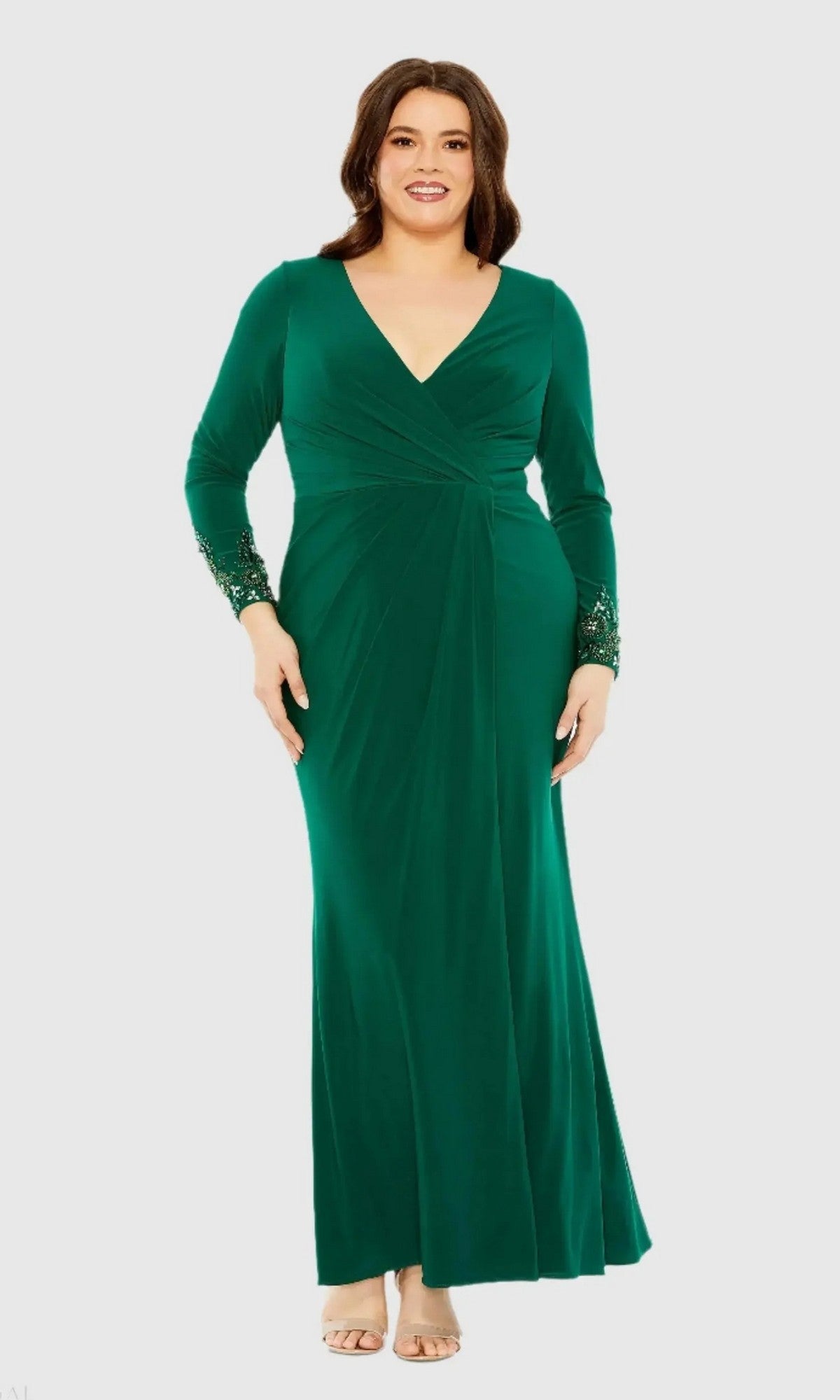 A woman wearing the Mac Duggal 77024 long plus-size formal dress in emerald, featuring long sleeves, a floor-length silhouette, a V-neckline, and beaded cuffs, stands against a plain light background.