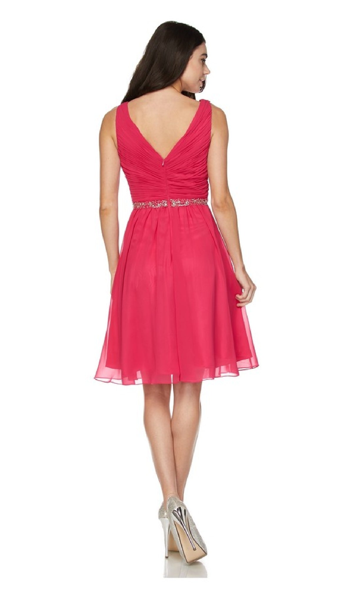 A woman models the Juliet 786 short semi-formal dress, featuring a sleeveless pink a-line design with a v-neckline, beaded empire waist, and V-back. She stands facing away, displaying silver high heels.