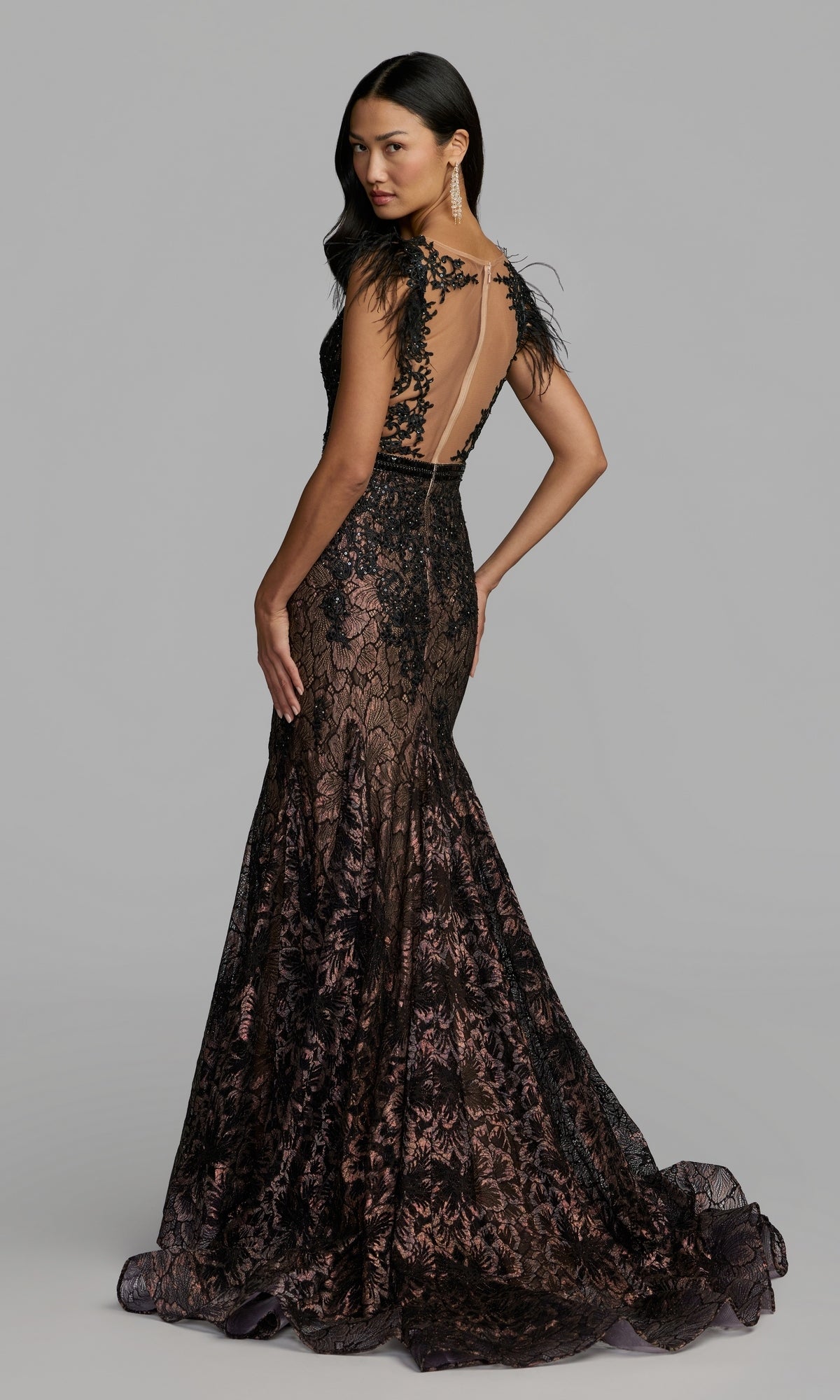 A woman wearing the Feathered Long Lace Formal Dress, Mac Duggal 79230, showcases its black and nude lace design with feathered cap sleeves, open back, and a floor-length mermaid silhouette while standing against a plain gray background.
