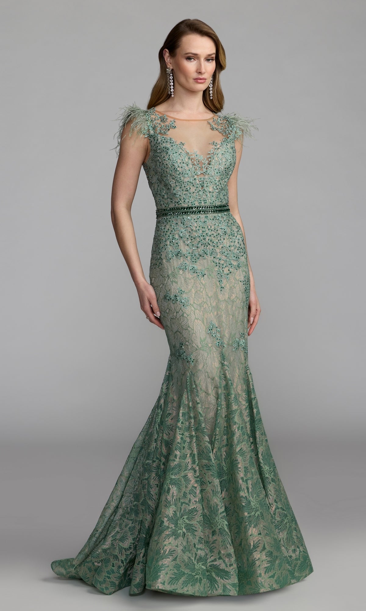 A woman is wearing the Mac Duggal 79230 Feathered Long Lace Formal Dress in sage—a floor-length, sleeveless gown with floral embroidery and feathered shoulder accents—paired with drop earrings, standing against a plain gray background.
