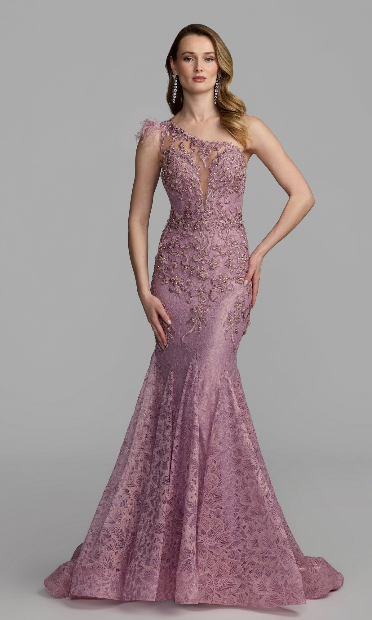 A woman stands wearing a fitted, floor-length rose gown—the Feather One-Shoulder Prom Gown: Mac Duggal 79264—adorned with elegant floral embellishments and featuring an eye-catching sheer panel at the neckline, set against a plain gray background.