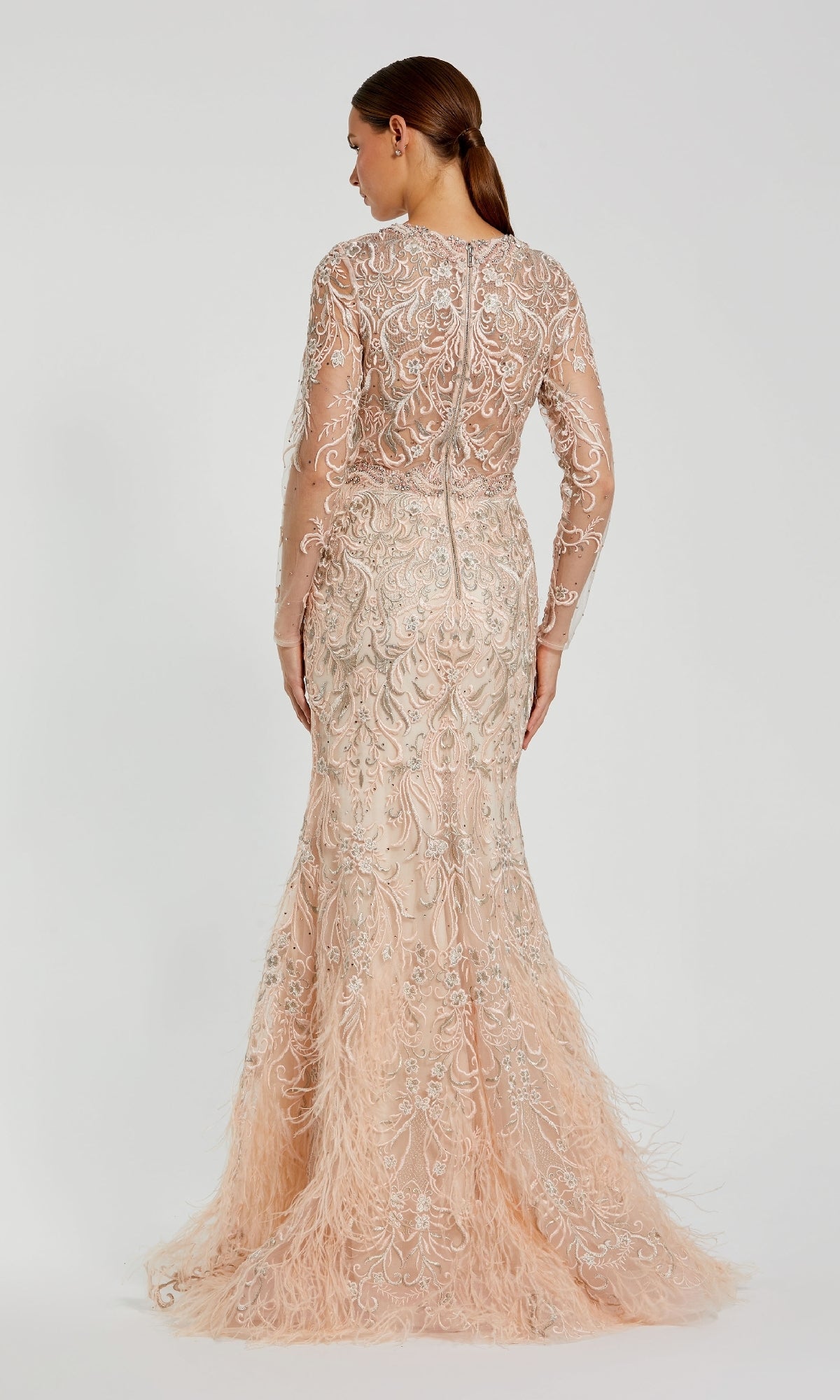A woman faces away, wearing the Mac Duggal 79267—a beaded long pink formal dress featuring intricate beadwork and feather details at the hem.
