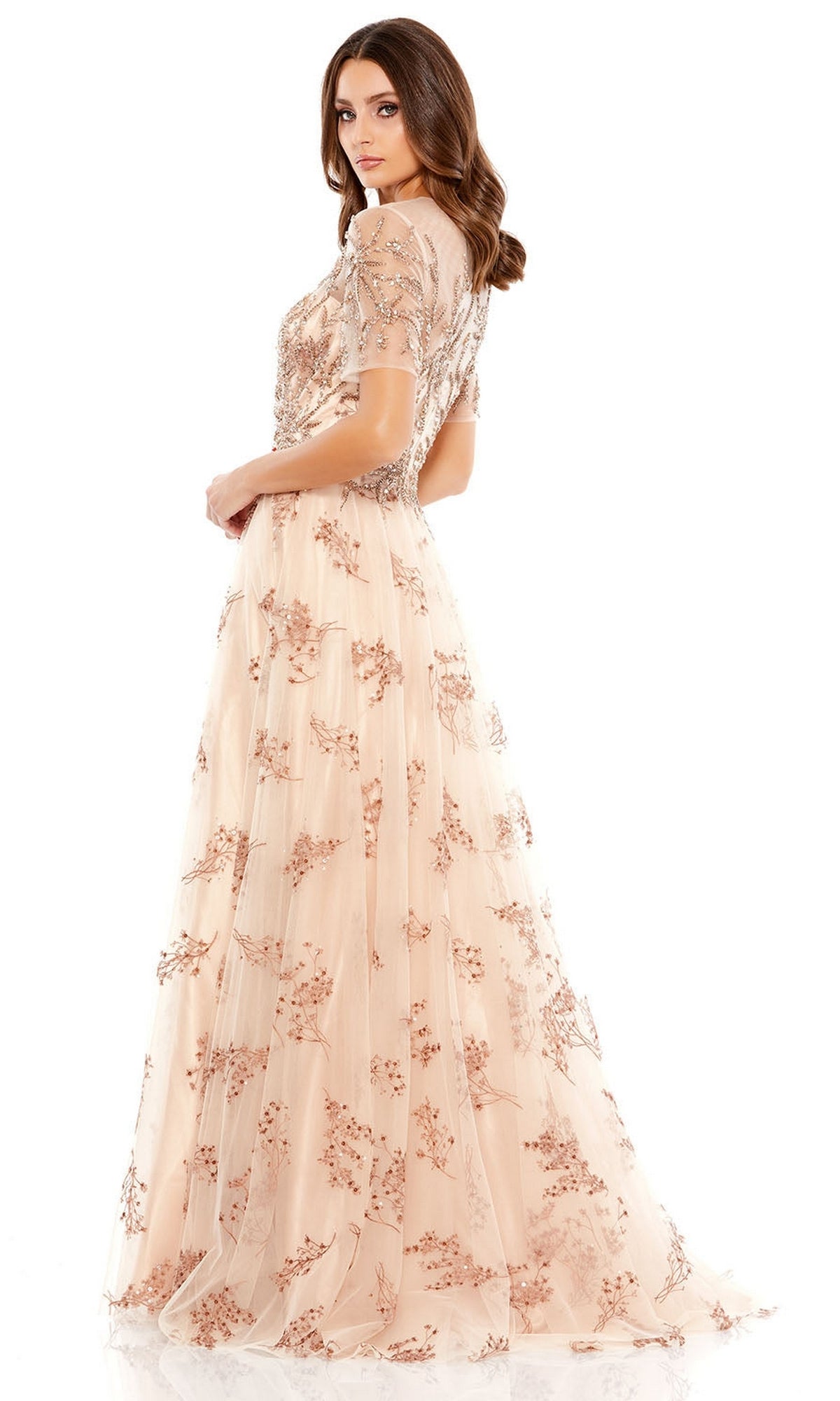 A woman stands looking over her shoulder, wearing the Long Formal Dress 79317 by Mac Duggal—a blush-colored gown with floral embellishments, short sleeves, and delicate embroidery.