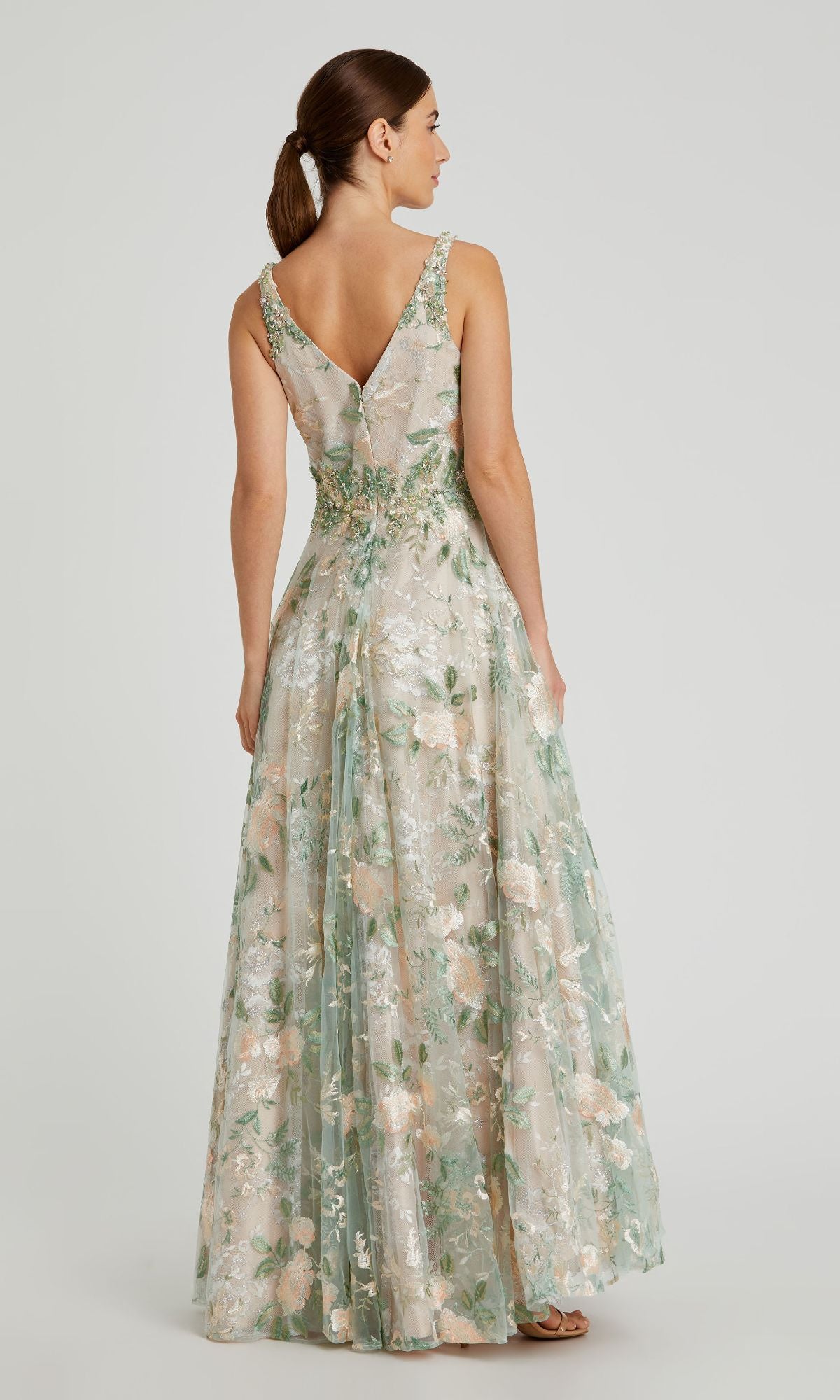 A woman faces away, wearing the Long Cream Floral Prom Dress Mac Duggal 79329 with a V-back and beige floral embroidery. Her hair is styled in a low ponytail.