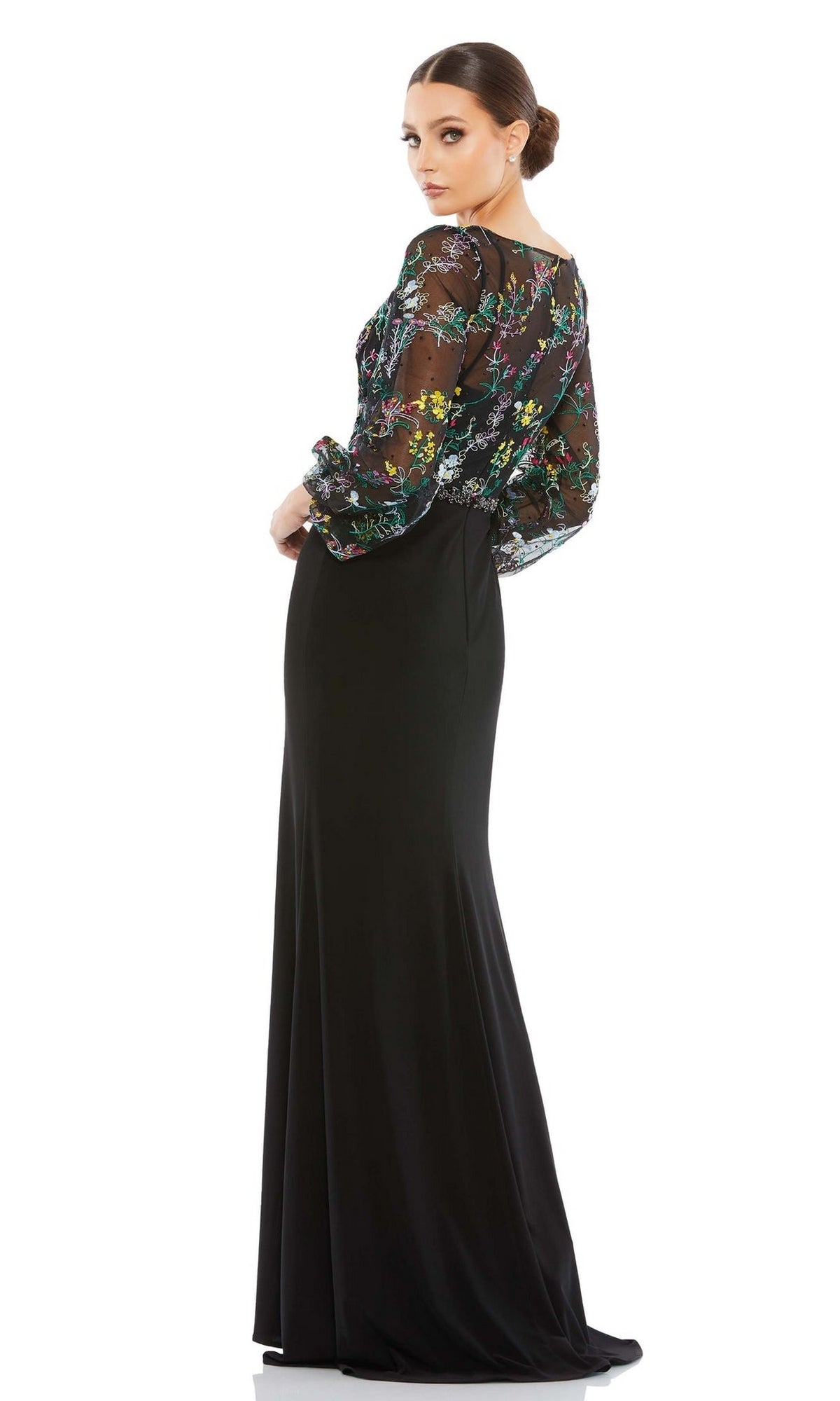 A woman stands in a low bun, looking over her shoulder, wearing the Mac Duggal 79379 long sleeve black formal dress with a sheer floral embroidered top and puffed sleeves.