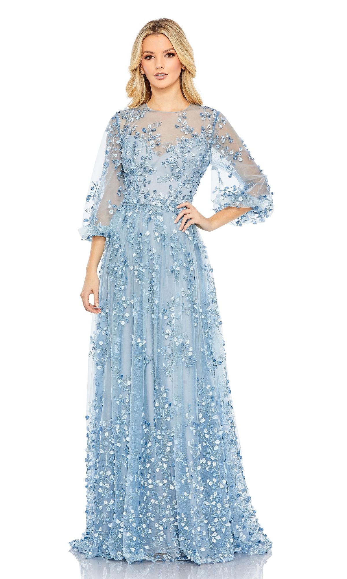 A woman wears the Mac Duggal 79399 long sleeve blue formal dress, featuring floral embroidery and sheer sleeves, against a white background.