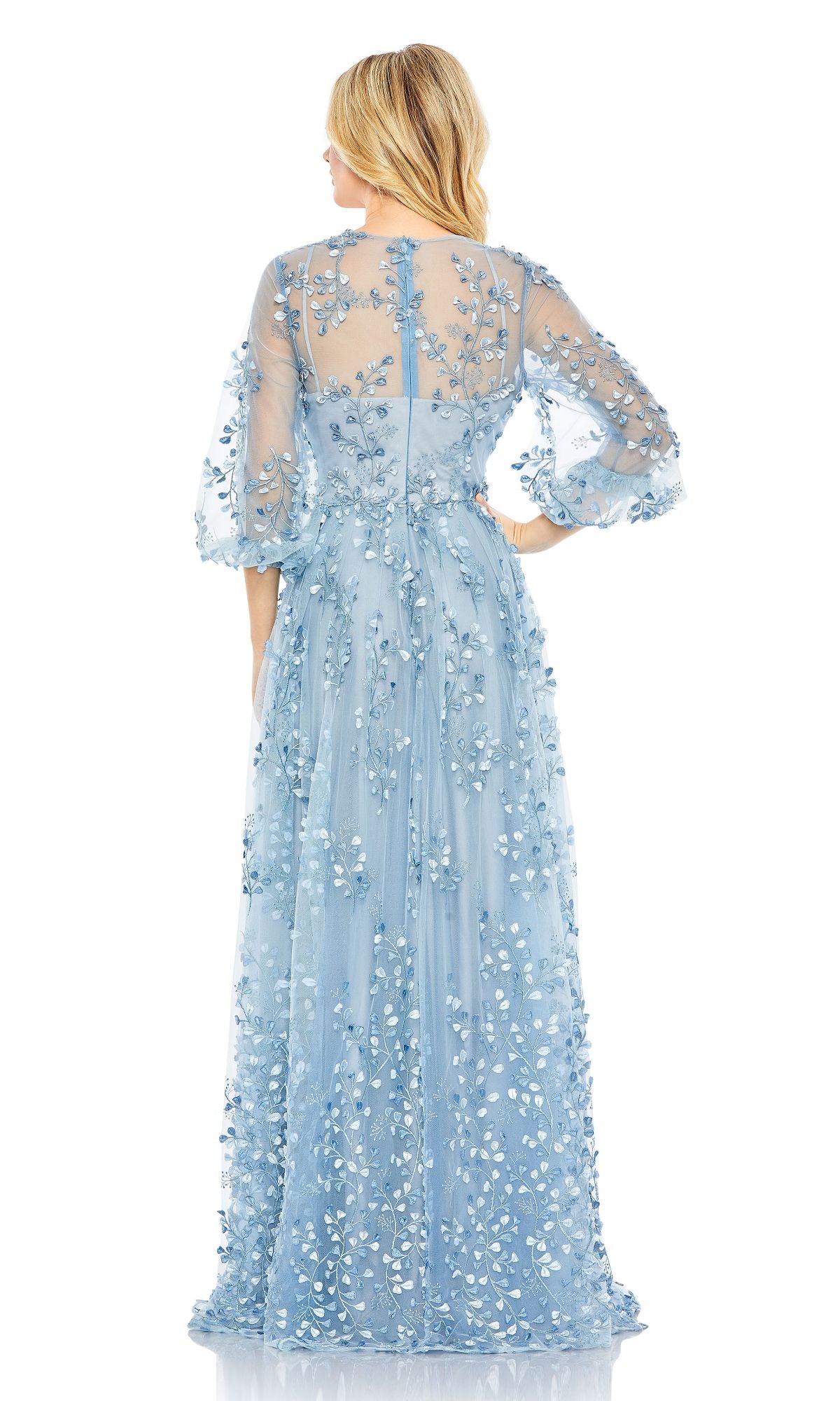 A woman stands facing away, wearing the Mac Duggal 79399 long sleeve blue formal dress featuring sheer sleeves and floral appliqué details.