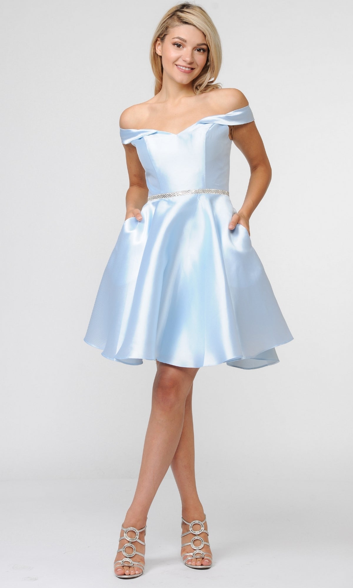 A woman models the Off-Shoulder Short Mikado Homecoming Dress 7948 in light blue, featuring a fitted bodice and flared skirt, paired with silver heels, against a plain background.