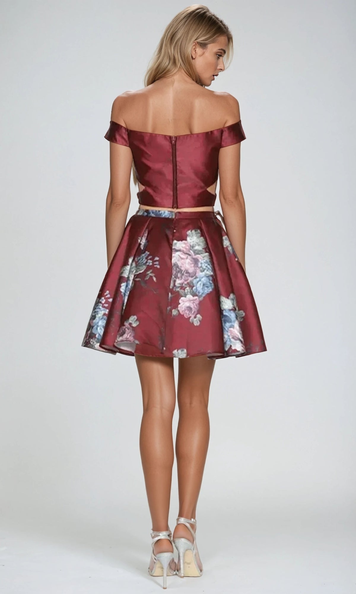 A woman faces away, wearing the Juliet 798 short party dress—an off-the-shoulder burgundy floral two-piece with an a-line skirt—paired with silver heels, set against a plain light background.
