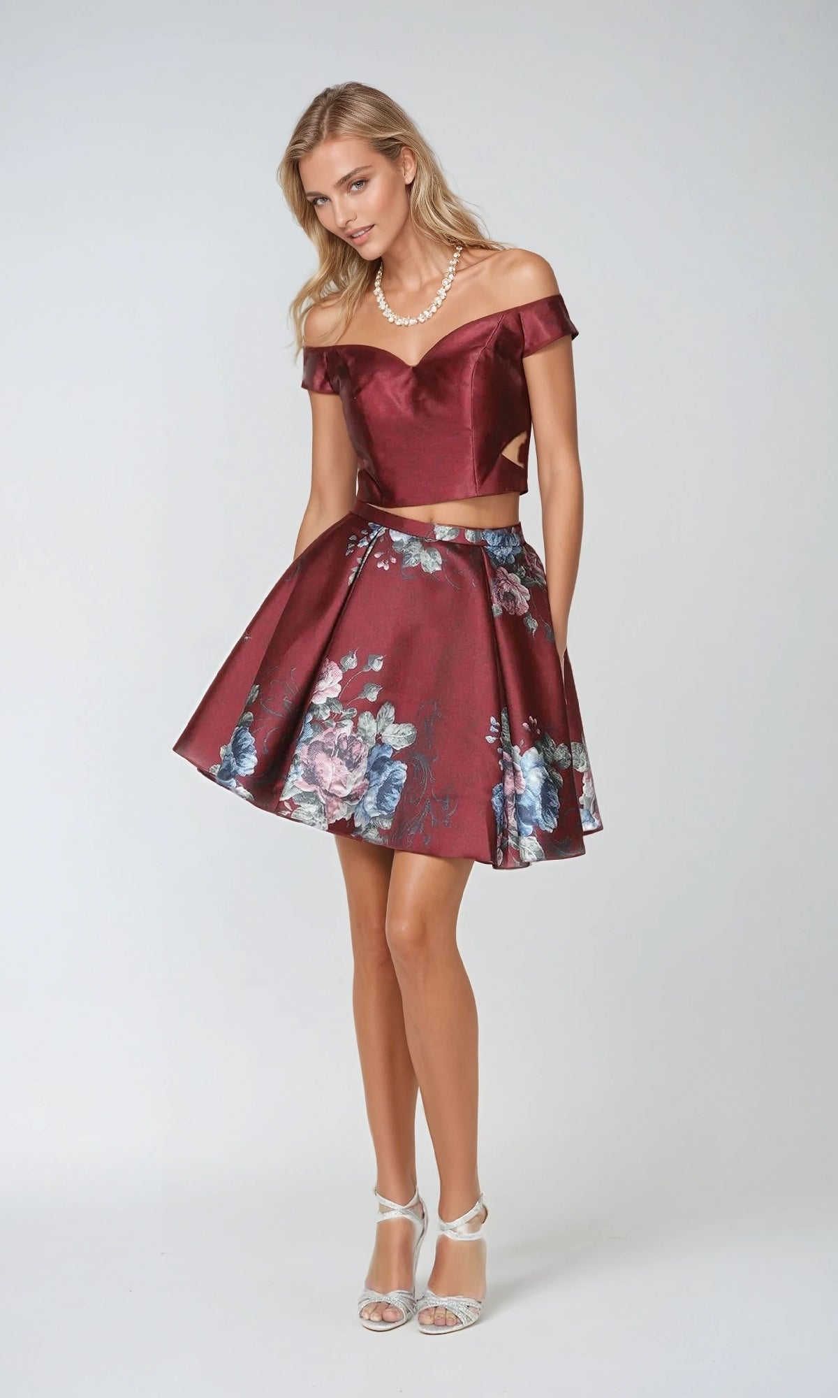 A woman in the Juliet 798 short party dress—burgundy off-shoulder top with a matching floral A-line skirt—stands against a light background, accessorized with a pearl necklace and white heels.