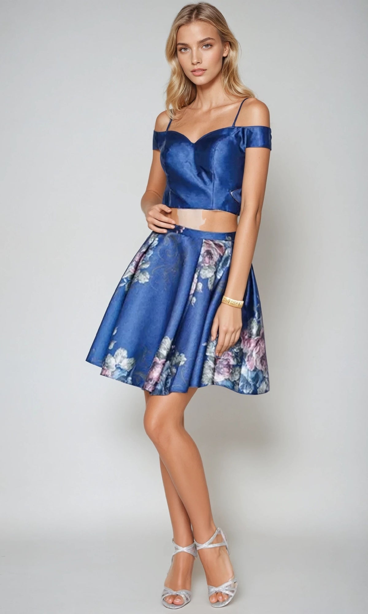A woman models the Juliet 798 short party dress, featuring a blue off-the-shoulder top and matching floral A-line skirt, styled with silver heels and a gold bracelet against a plain background.