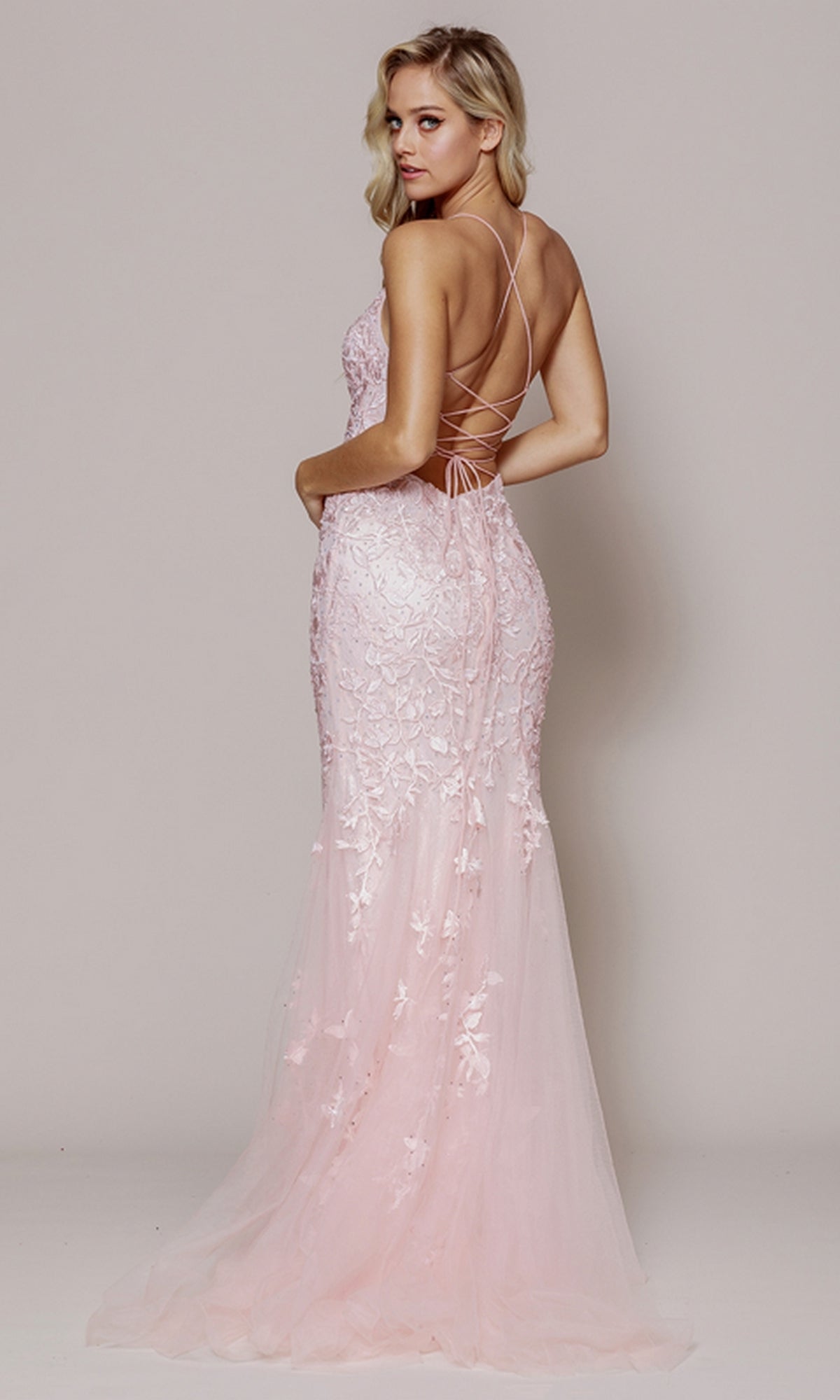A woman stands looking over her shoulder in the Embroidered-Lace Tight Long Prom Gown 799—a light pink, floor-length evening dress with a fitted bodice, delicate embroidery, and crisscross straps on an open back.