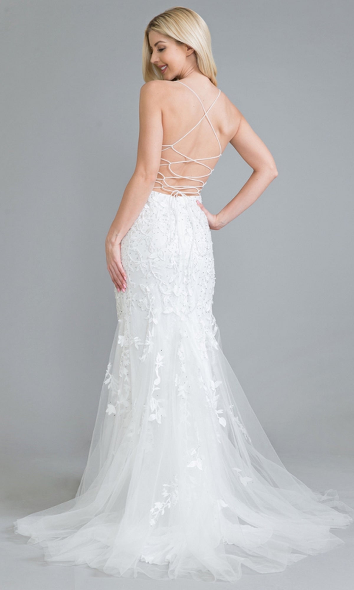 A woman models the Embroidered-Lace Tight Long Prom Gown 799, featuring a backless crisscross design, sheer train, and intricate embroidery, standing against a plain gray background.