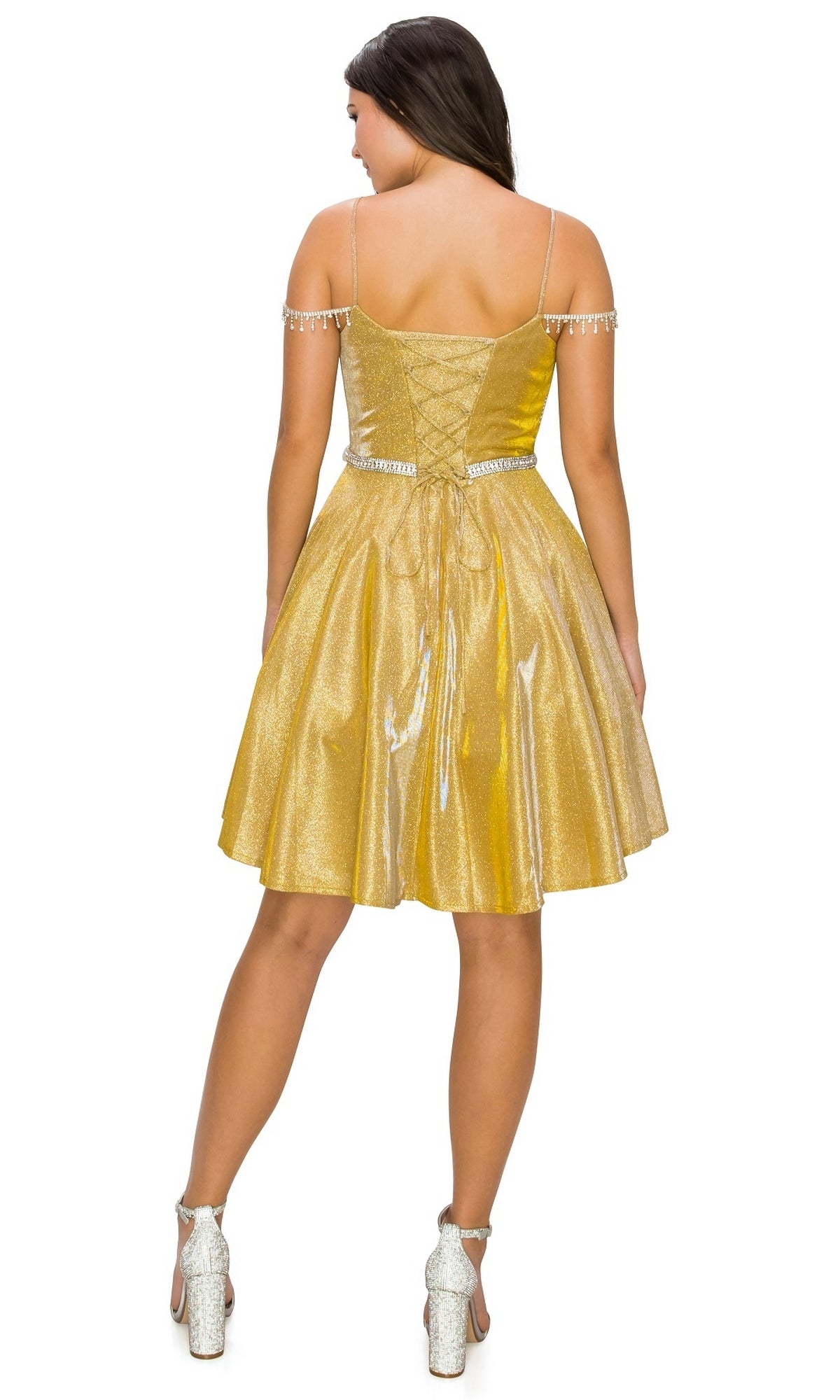 A woman stands with her back to the camera, wearing the Beaded Cold-Shoulder Short Homecoming Dress 8014J in gold metallic shimmer with decorative straps and silver heels—a chic choice for any semi-formal event.